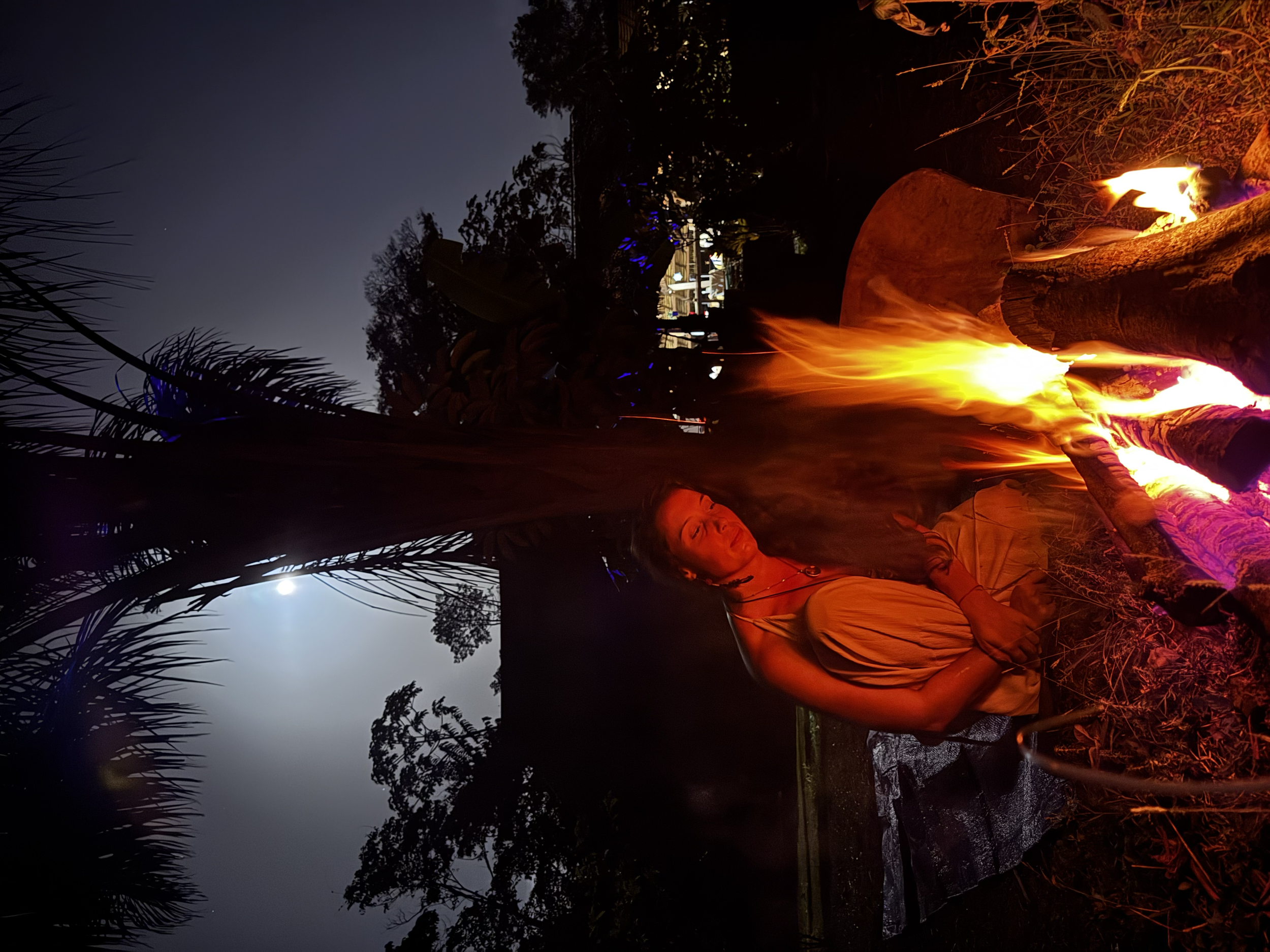Une femme assise près d'un feu de camp en soirée, avec la lune et des arbres en arrière-plan.