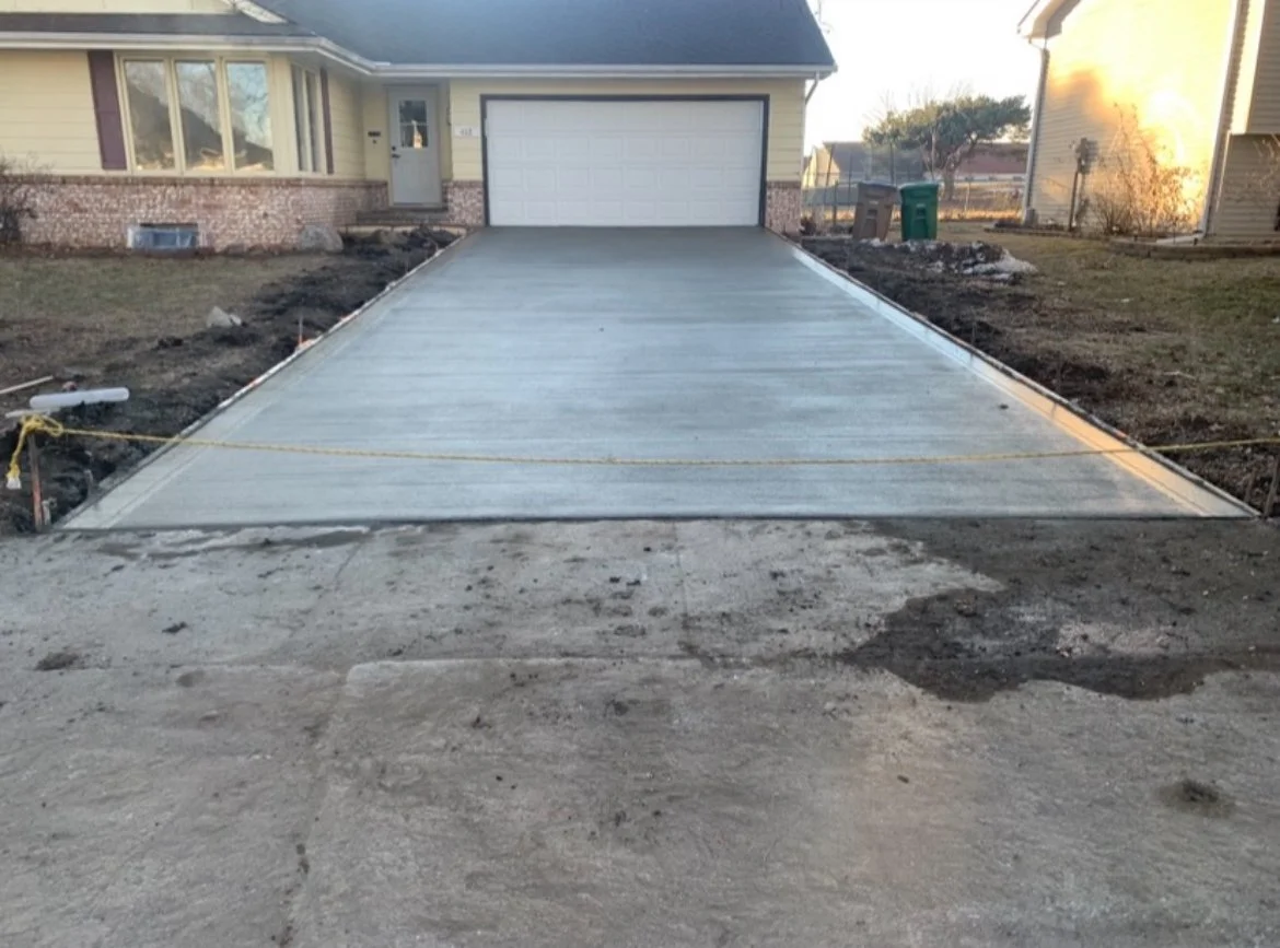 Newly constructed concrete driveway leading to a garage of a house, with some construction materials and soil on the sides.