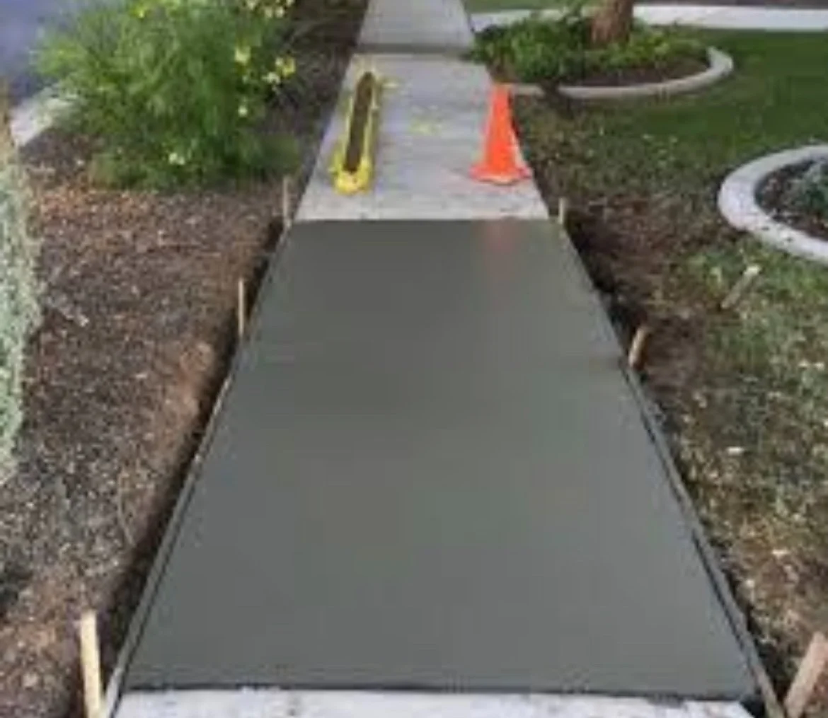 Newly poured concrete sidewalk under construction, with a level and orange traffic cone on a residential lawn.