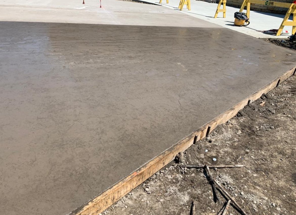 Freshly poured concrete on a sidewalk under construction, with construction tools and barriers in the background.