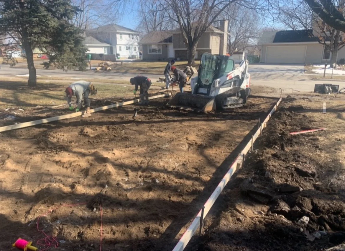Construction workers are working on a residential yard, leveling the ground with a skid steer and wooden framing for a new sidewalk or foundation.