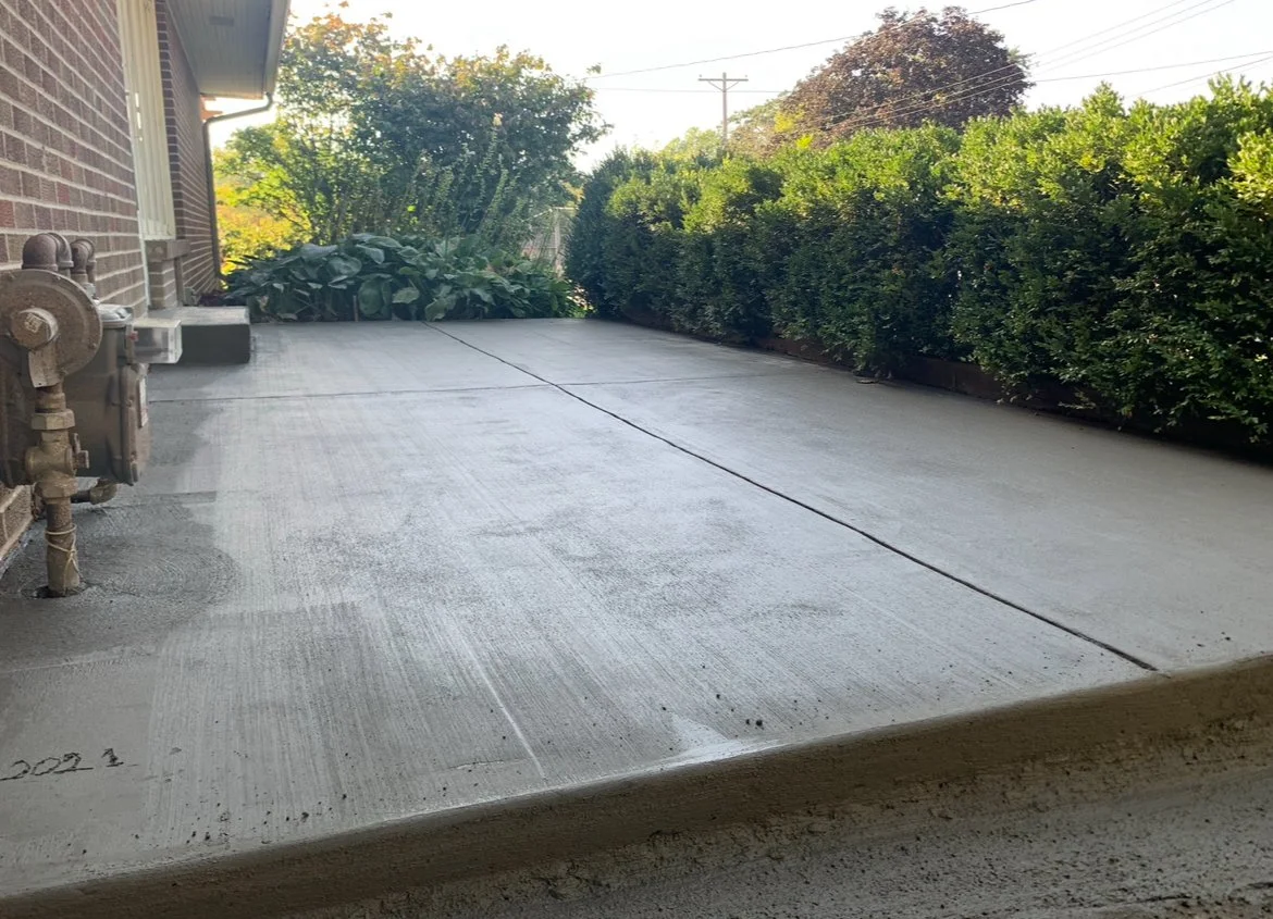 Newly poured concrete patio with bushes on the right side and a brick house wall on the left, under clear sky.