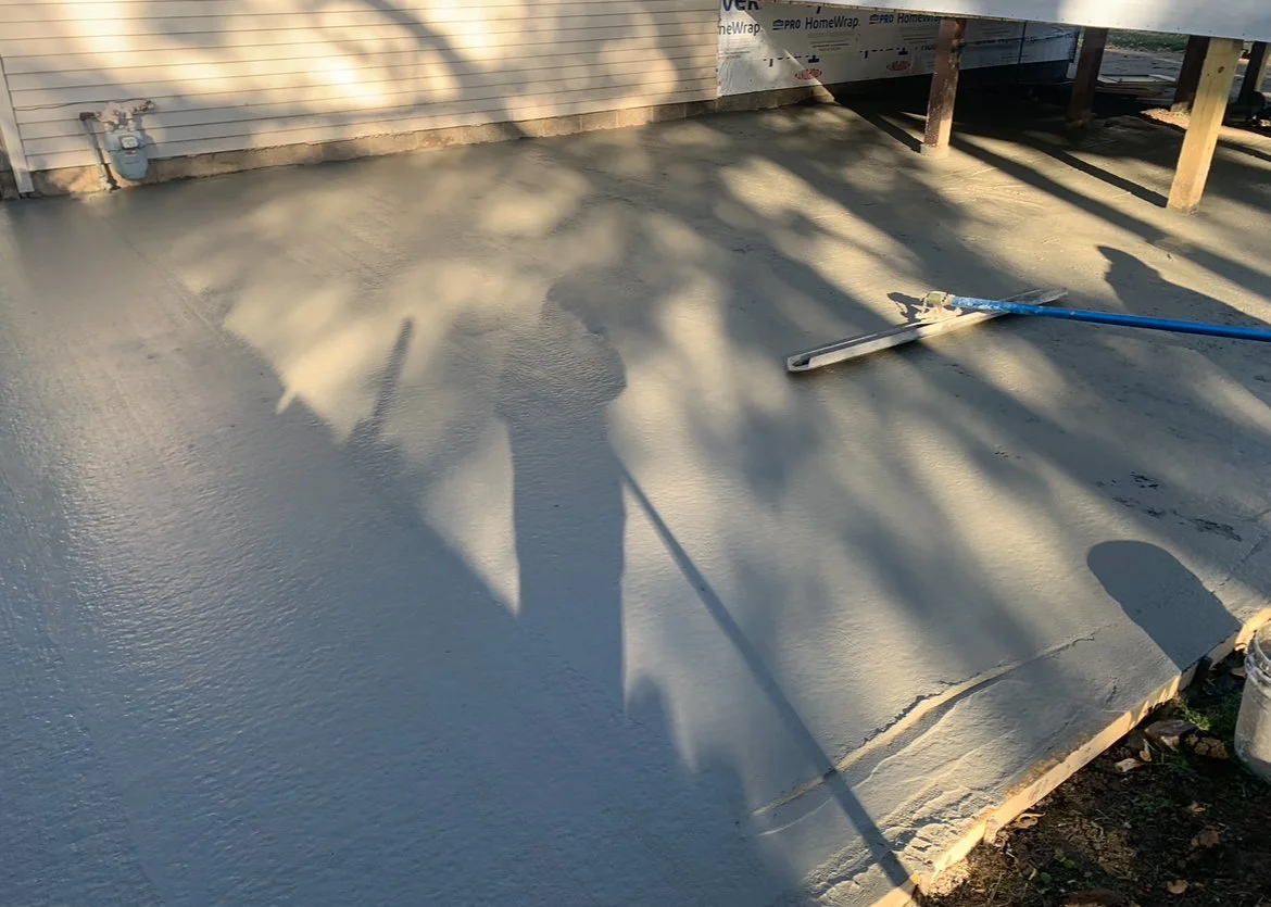 Freshly poured concrete on a building's porch or sidewalk with construction tools and shadows of trees.