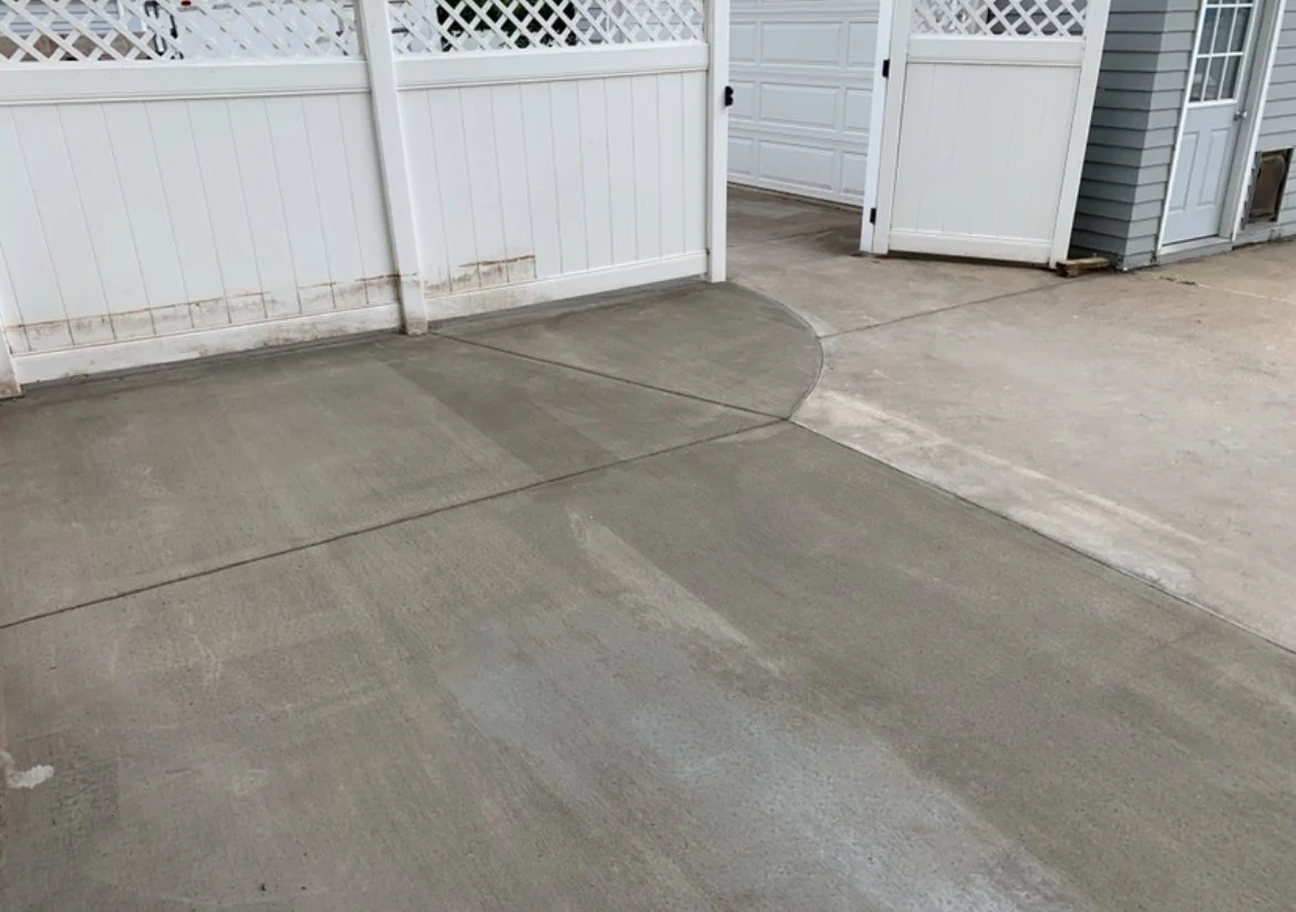 Freshly poured concrete driveway with a curved edge near a white fence and a garage door.
