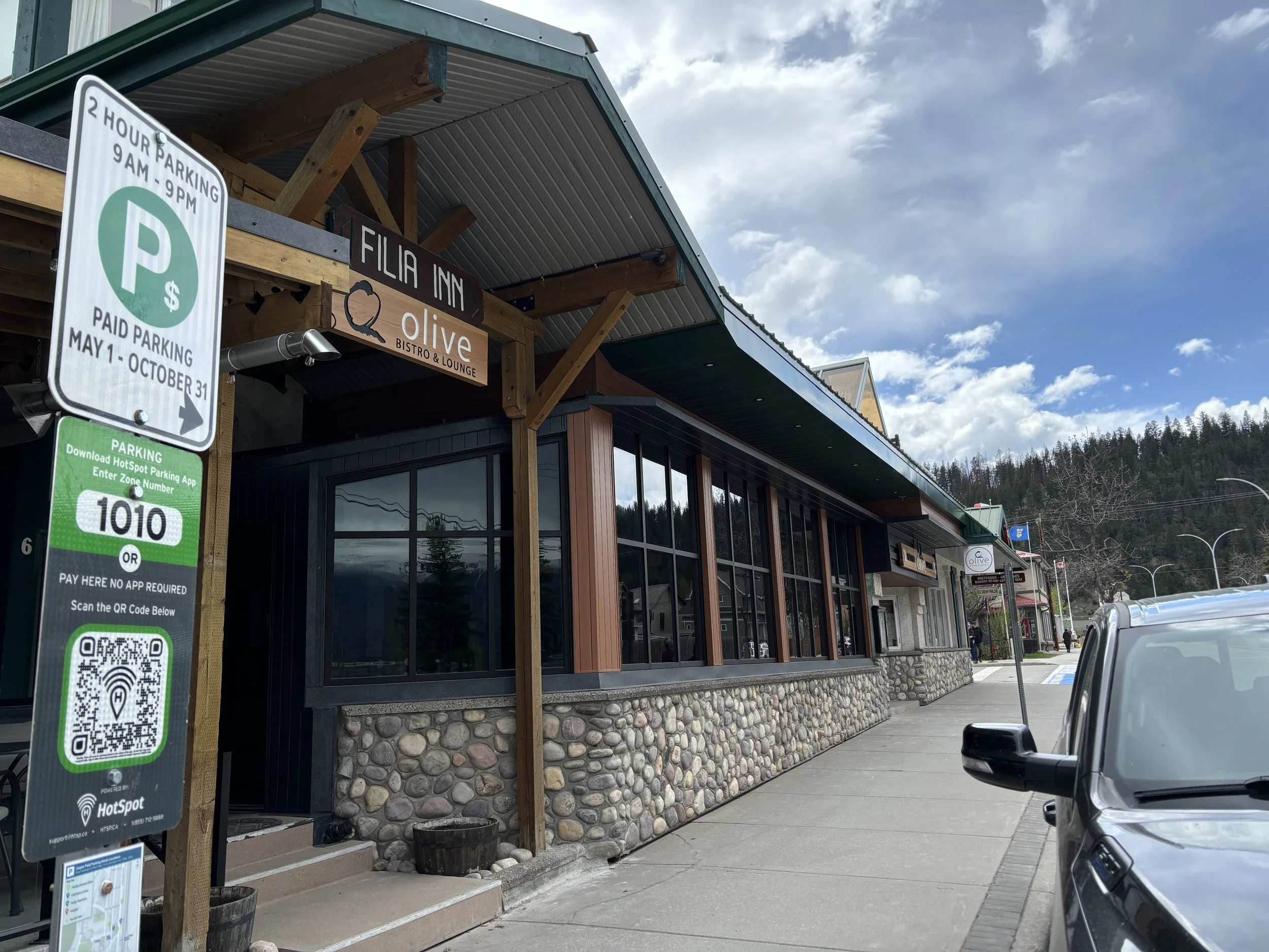 Street view of a restaurant named Olive Bistro & Lounge with large windows, stone facade, and parking signs in front, with a partially cloudy sky and hills in the background.