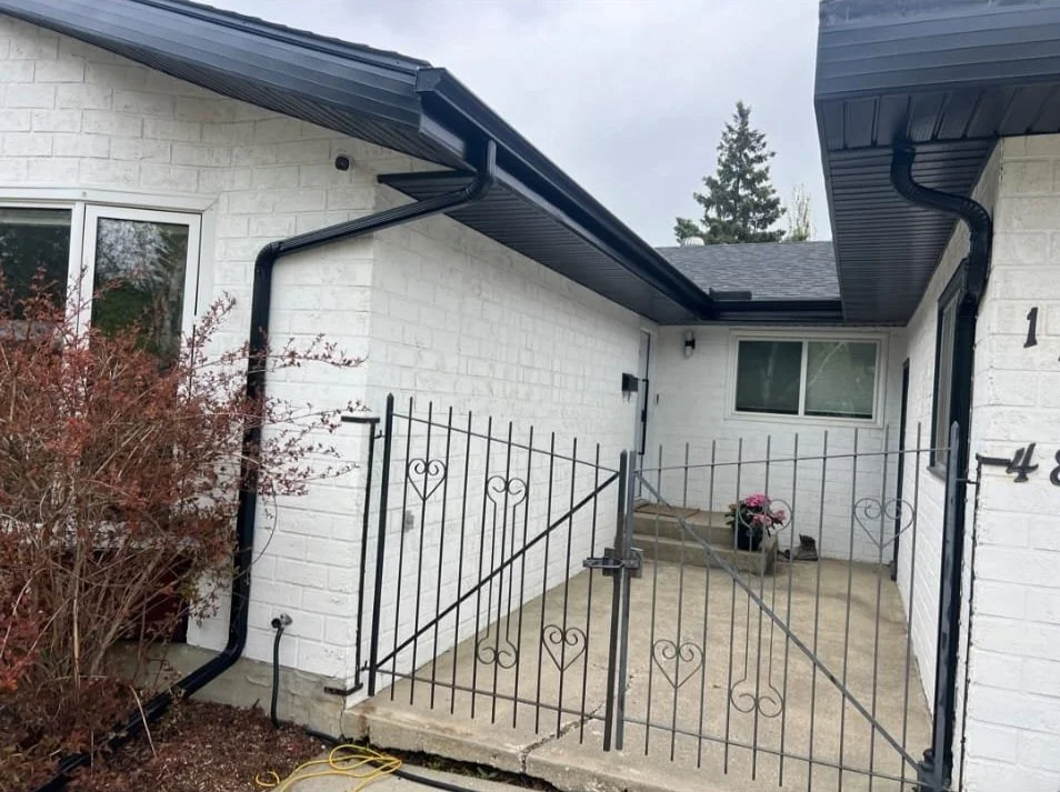 Front entrance of a house with a white brick wall, black metal gate, and black gutters. There are plants and a window visible.