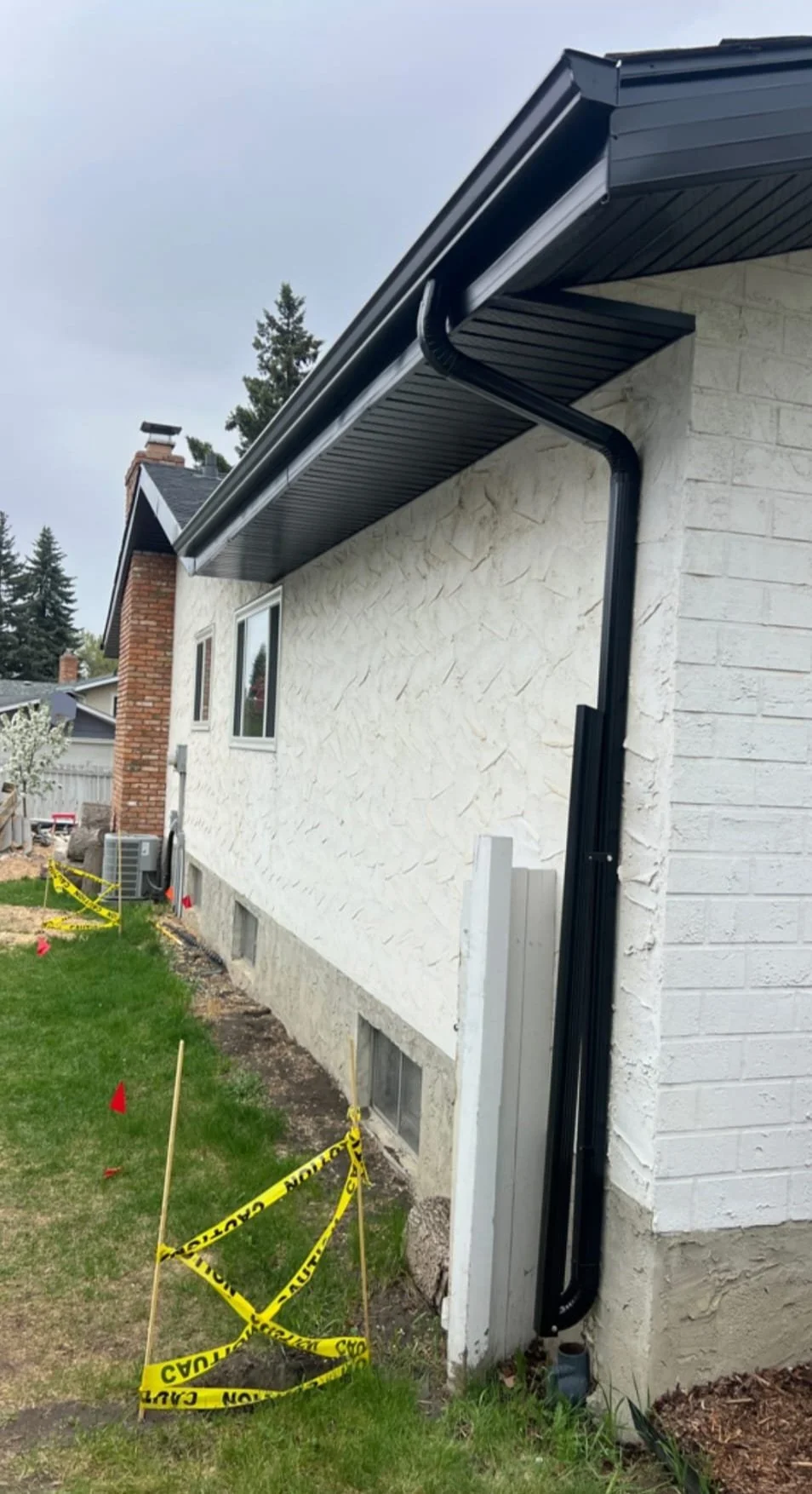 The side of a house with newly installed black gutter and downspout system, in front of the house is a small lawn with caution tape and small flags marking work area.