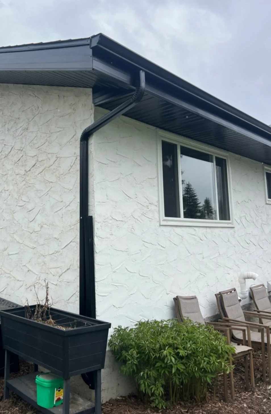 The exterior of a house with a white textured wall, a black gutter system, and several garden chairs. There is a large window reflecting trees and sky, along with a green bush and a planter with dried plants.