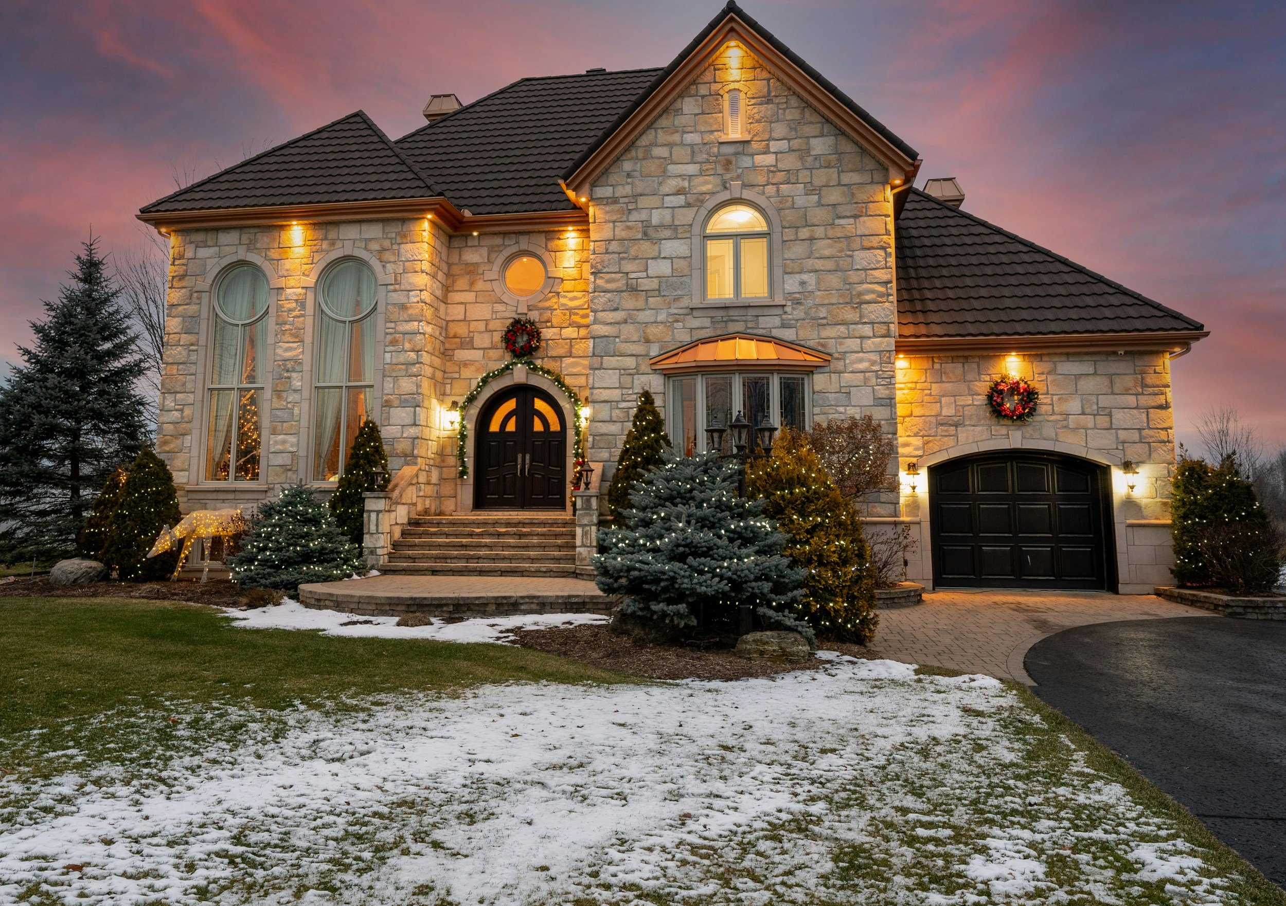 A large stone house decorated for Christmas with wreaths, lights, and trees, illuminated at dusk, with a backyard with patches of snow.