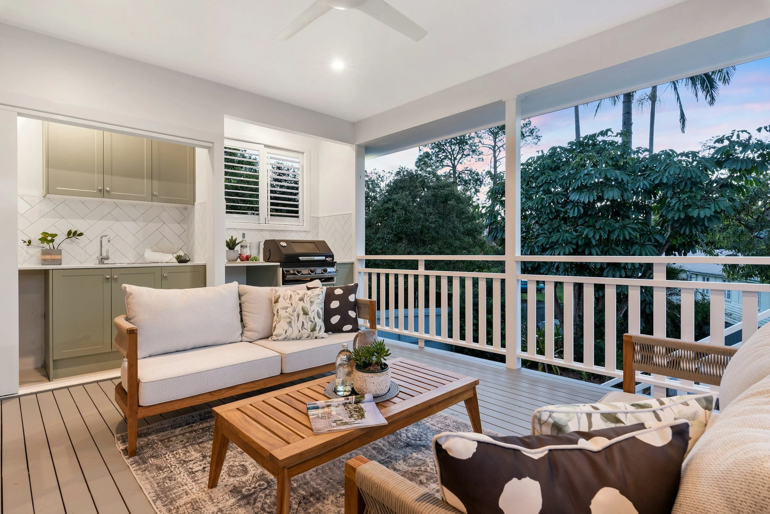 An outdoor balcony with a seating area, including a white couch with patterned pillows, a wooden coffee table with magazines, and a patterned rug. The balcony has a railing, a small kitchen area with beige cabinets, a grill, and large windows. Lush green trees and a colorful sky are visible outside.