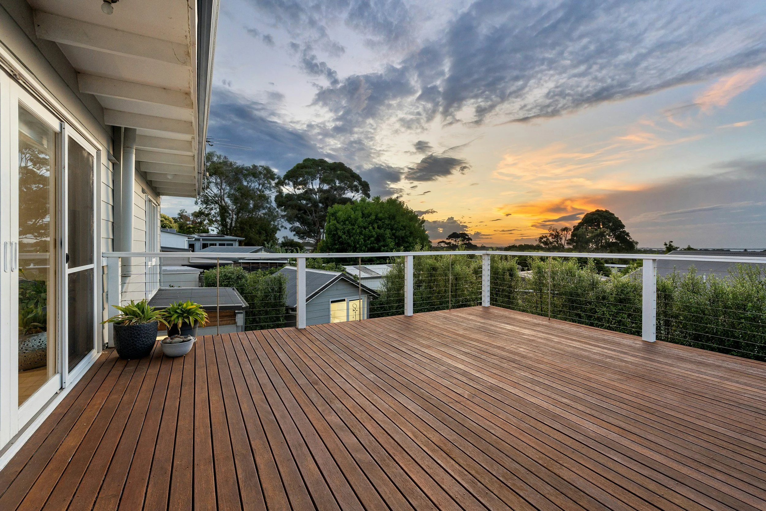 A spacious wooden deck attached to a house, with two potted plants near glass sliding doors on the left, overlooking a neighborhood with rooftops and tall trees under a partly cloudy sky at sunset.