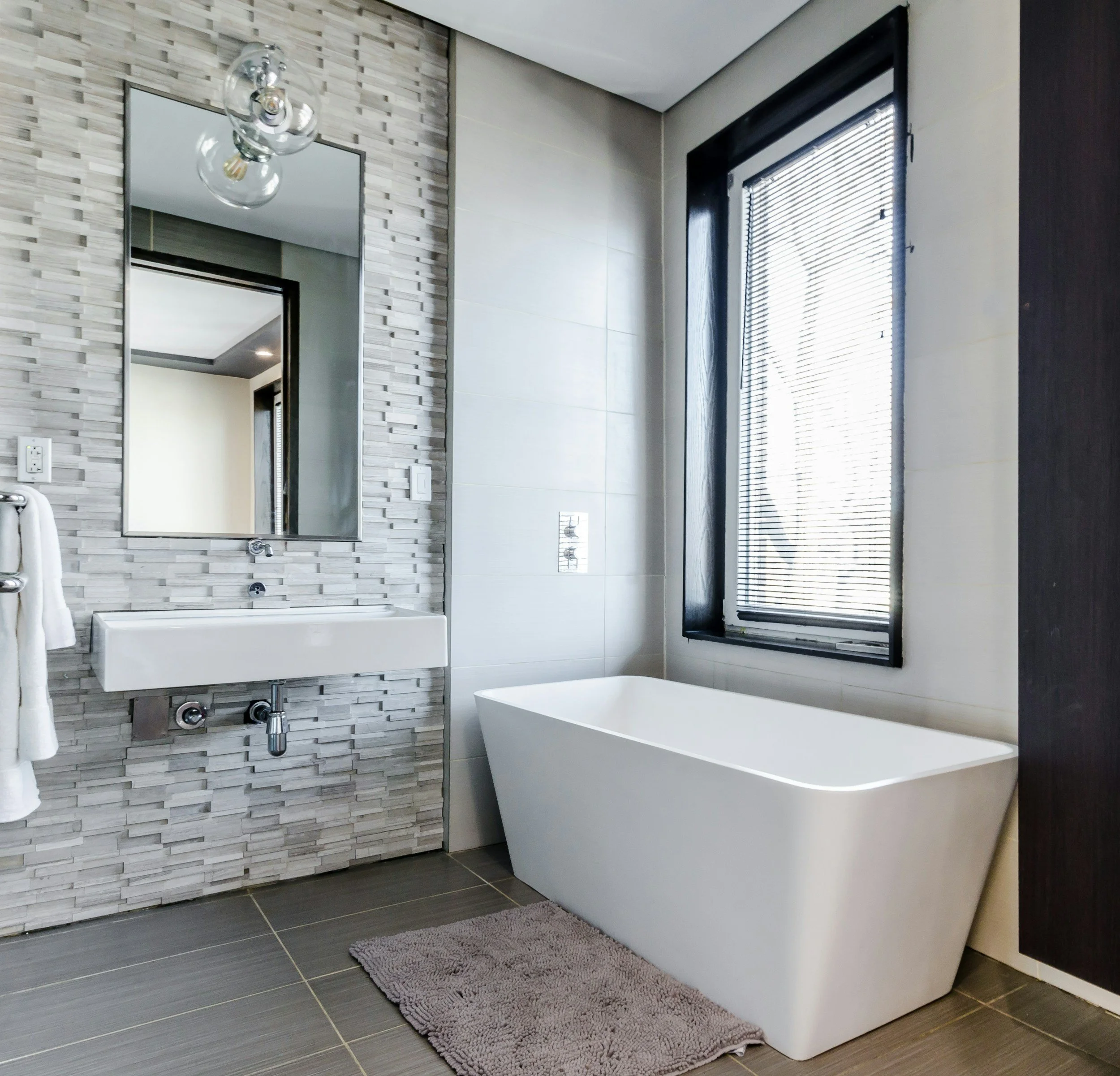 Modern bathroom with gray tile floors, a white freestanding bathtub, a window with black blinds, a textured tile wall, and a wall-mounted mirror.