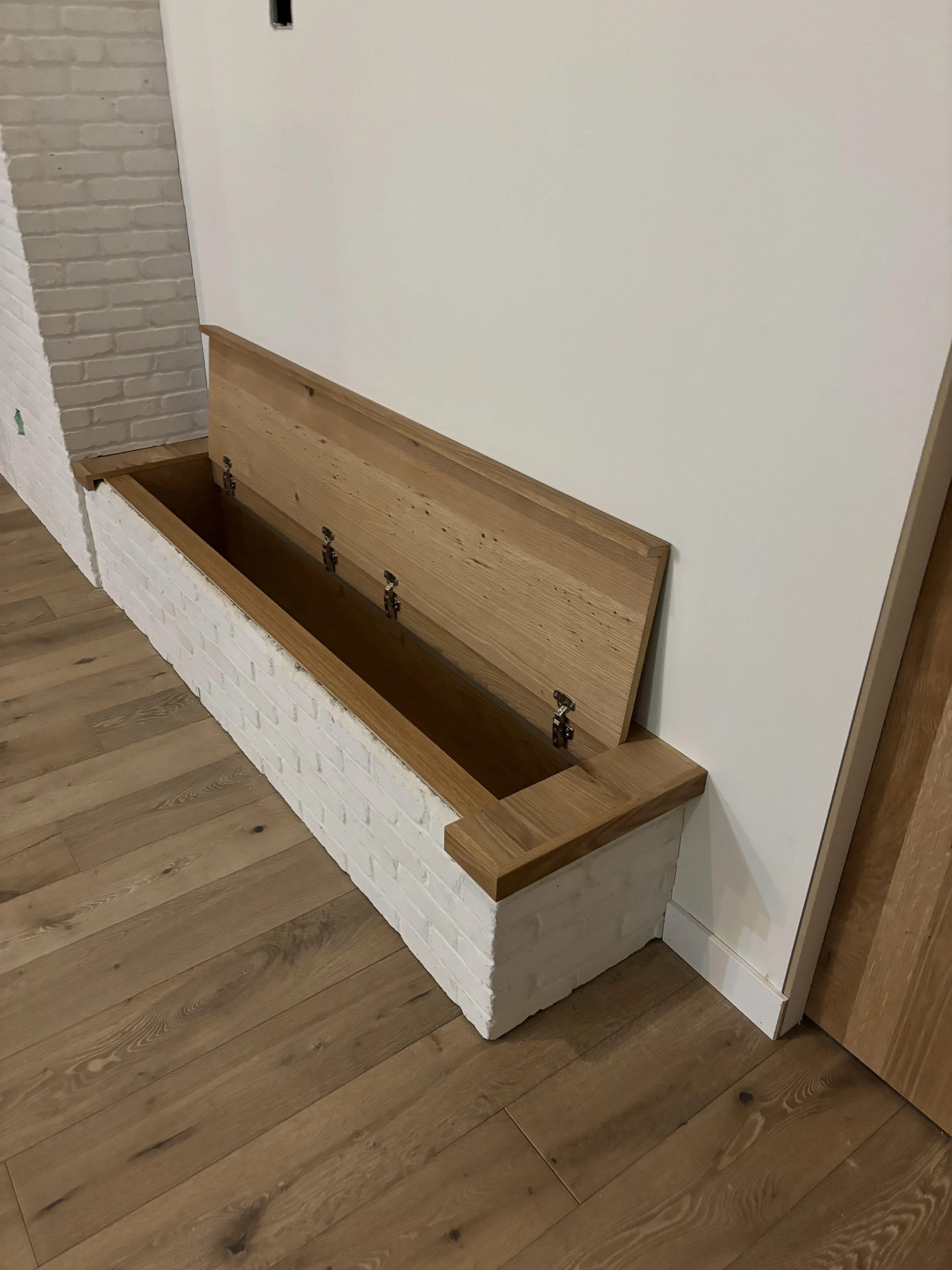 Built-in wooden storage box with hinged lid, set into a white brick base along a wall, with a wooden floor nearby.