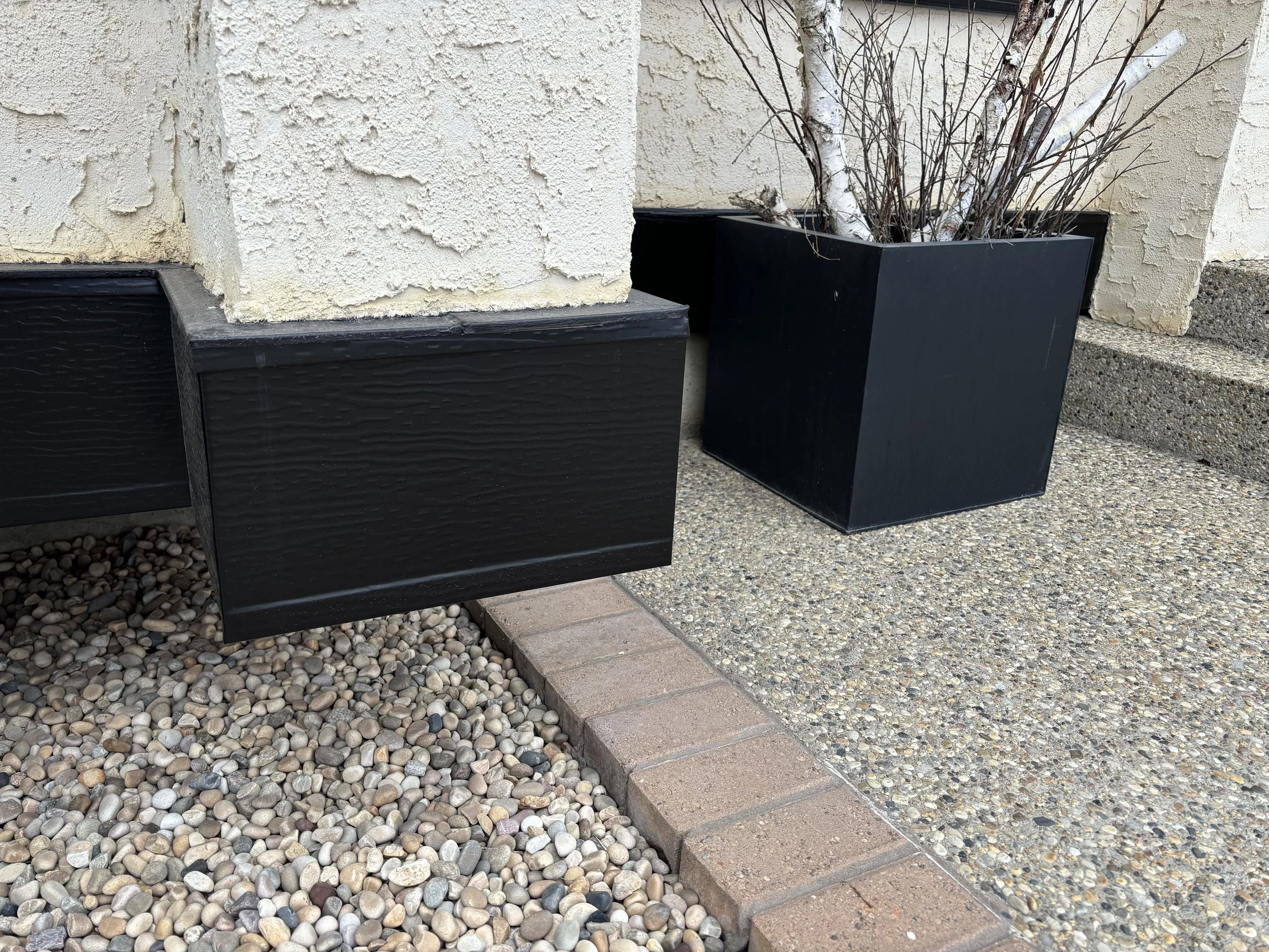 Black rectangular plant boxes with a tree and some dry branches outside on a concrete and pebble ground near a building wall.