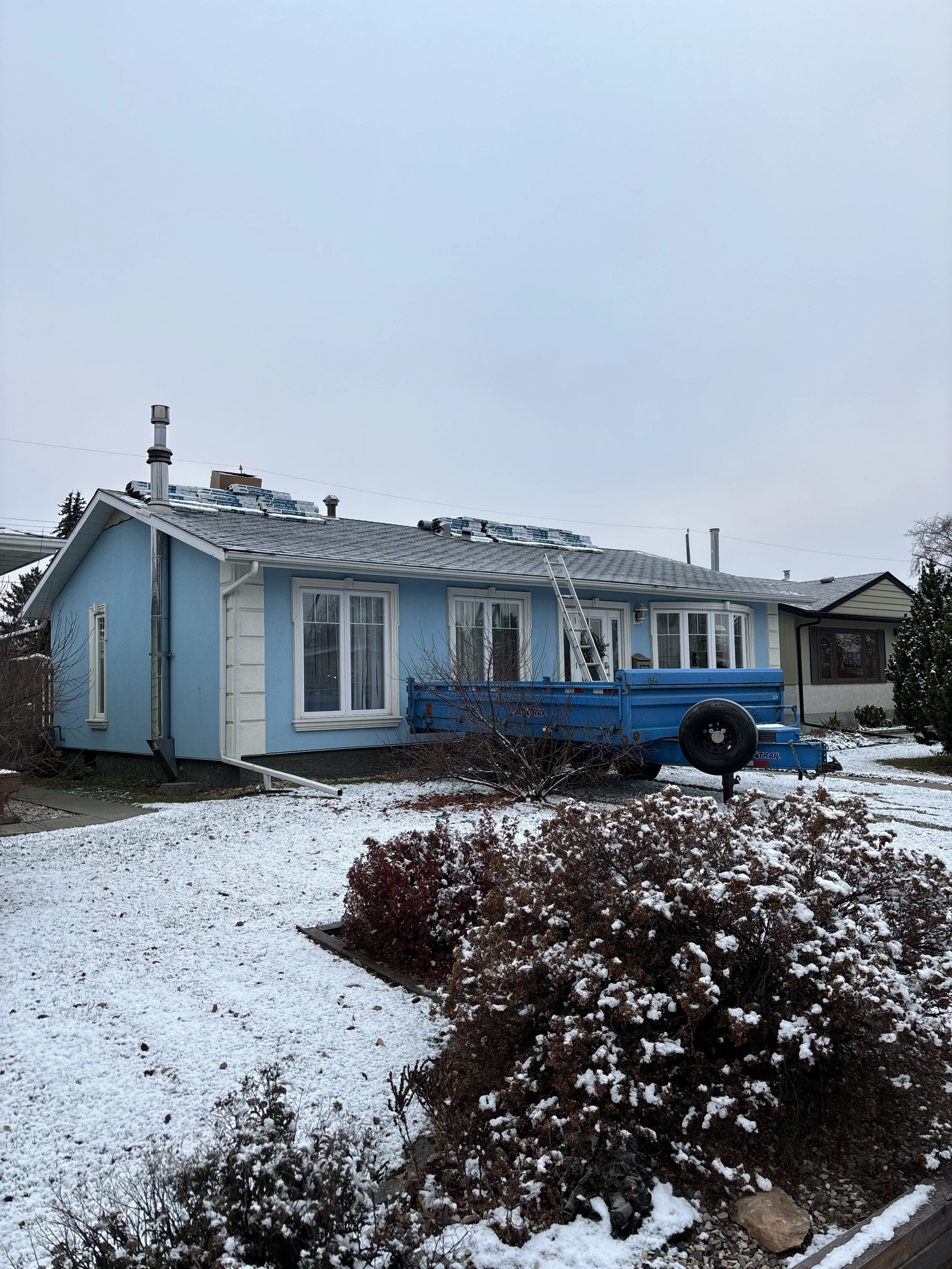 A blue house with snow on the roof, a trailer parked in front, and a ladder leaning against the house during winter.