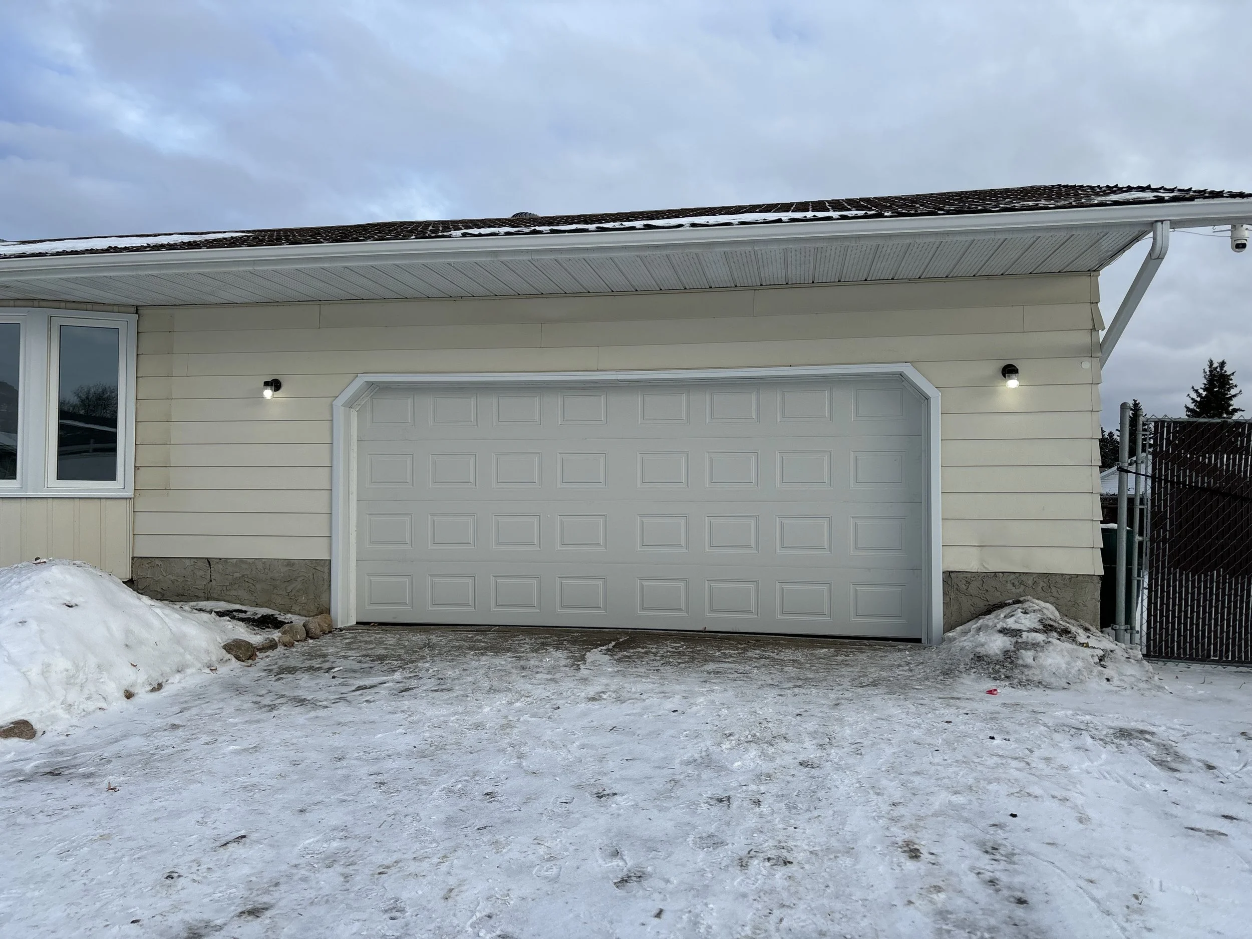 Exterior view of a house with a closed white garage door, snow-covered driveway, and two outdoor lights on either side of the garage door