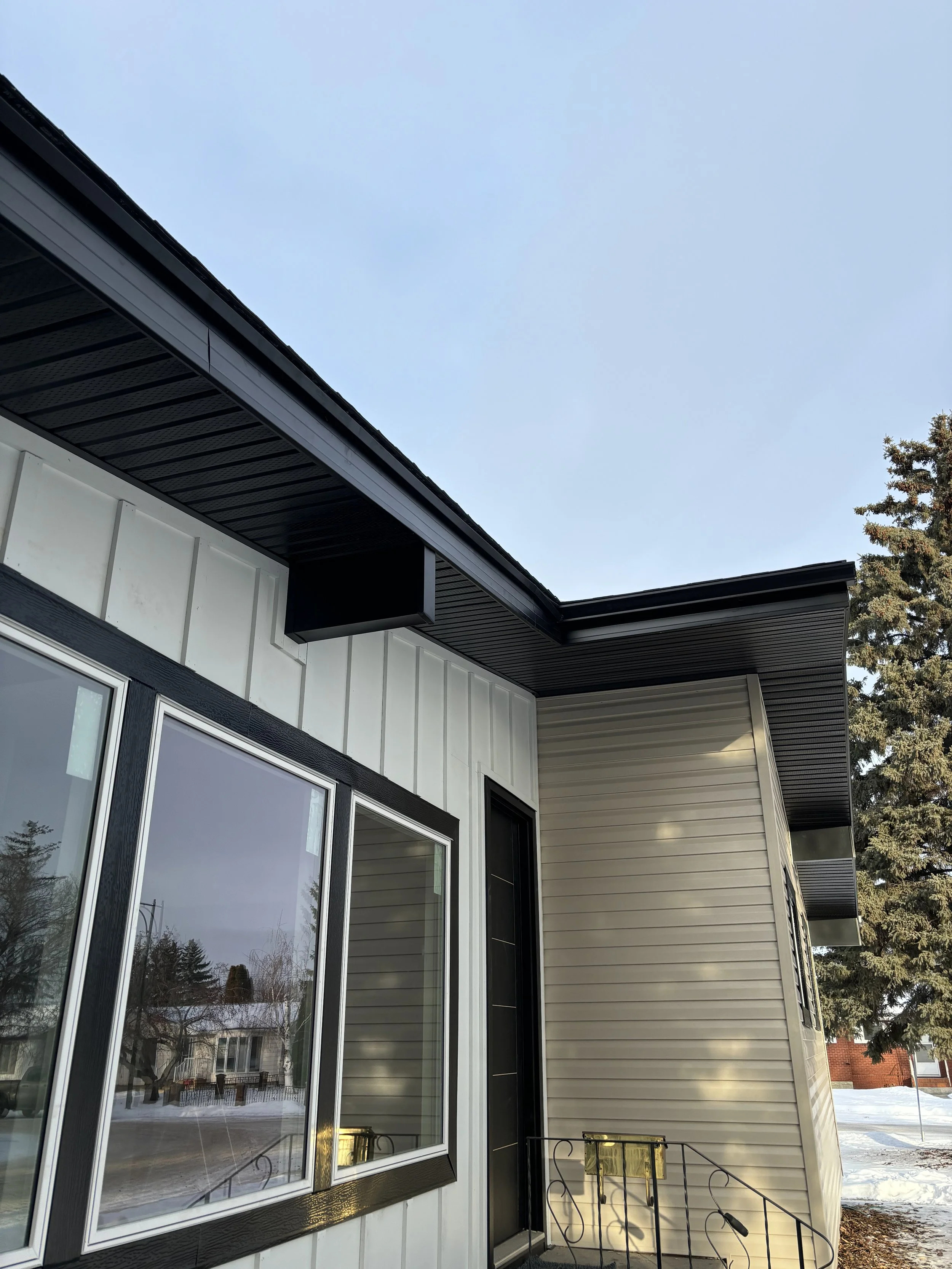 Close-up of the exterior corner of a modern house with white and beige siding, large window, black trim, and metal staircase railing, during winter with snow on the ground.