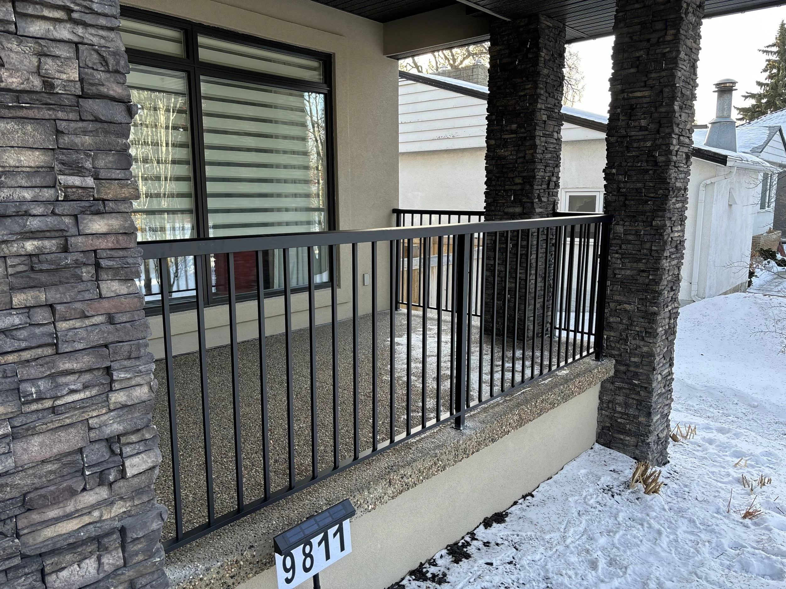 A balcony with a black metal railing, stone columns, and a stucco wall, with snow on the ground and a residential neighborhood in the background.