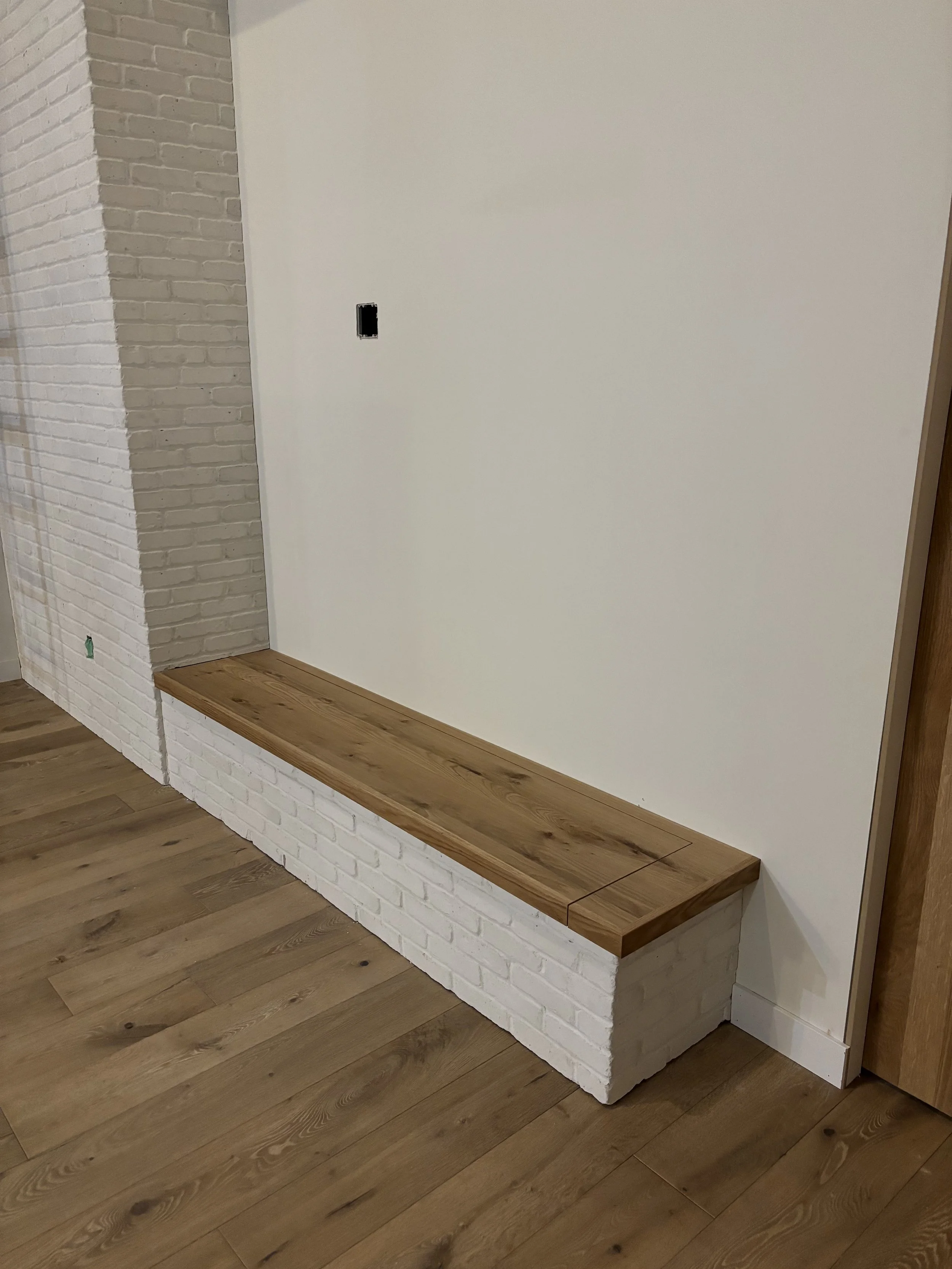 Indoor wall with white brick base and a wooden bench or ledge, white wall, electrical outlet, and floor with wood planks.