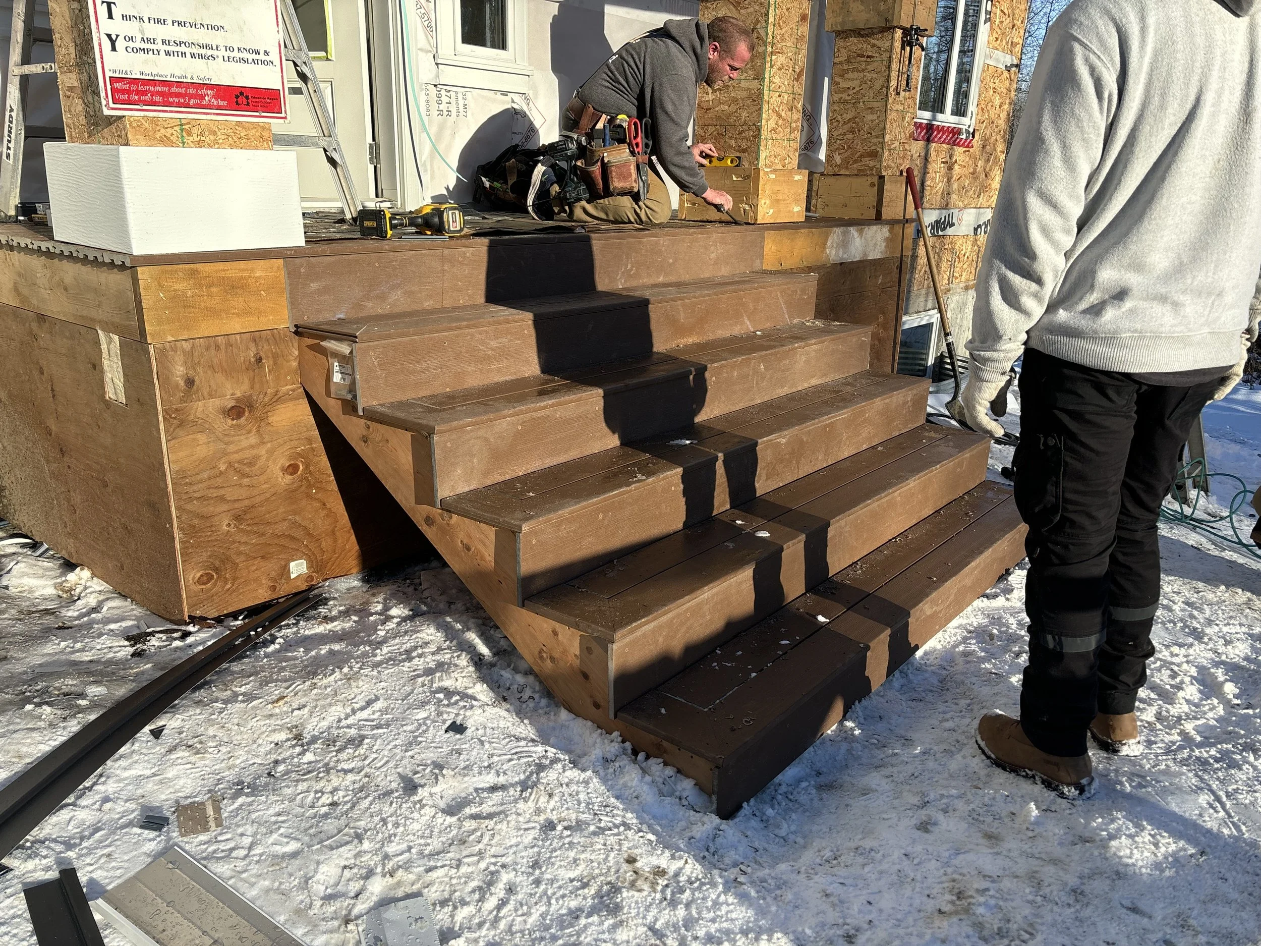 Construction workers installing wooden stairs outside a house on snow-covered ground.