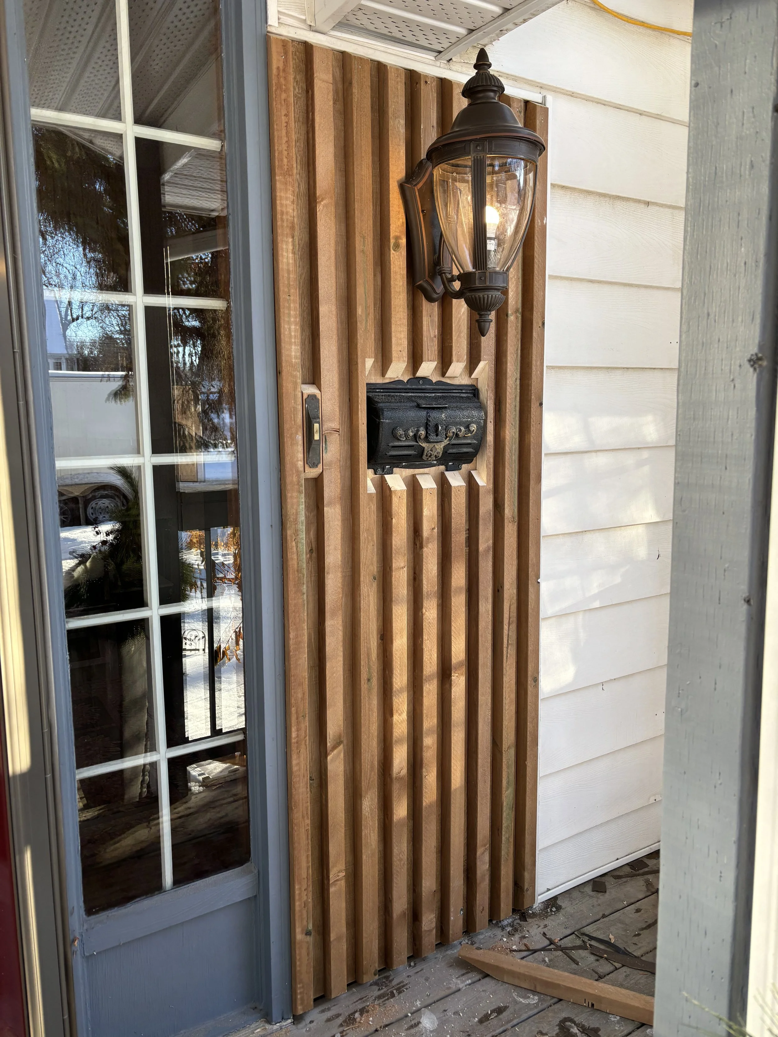 A front porch with a black outdoor lamp, a mailbox, wooden siding, and part of a glass double door.