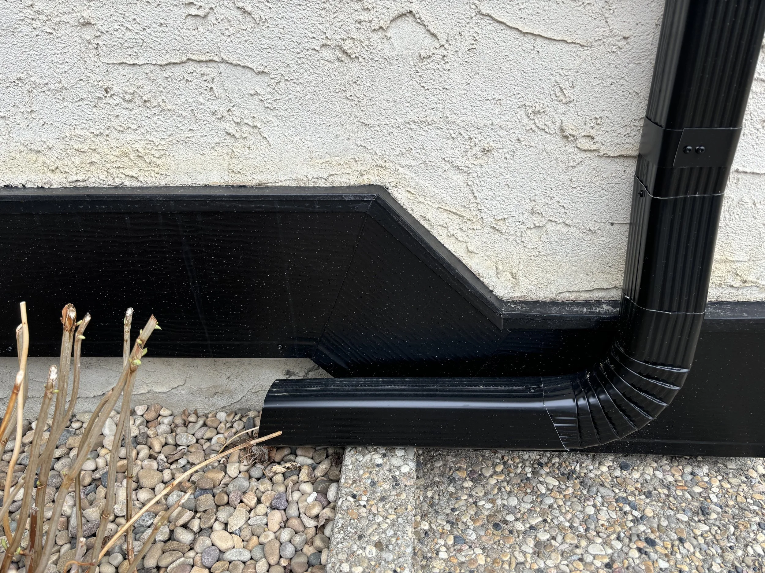 Black drainage system with a downspout connected to a gutter along the side of a house, with gravel and some dried plants nearby.