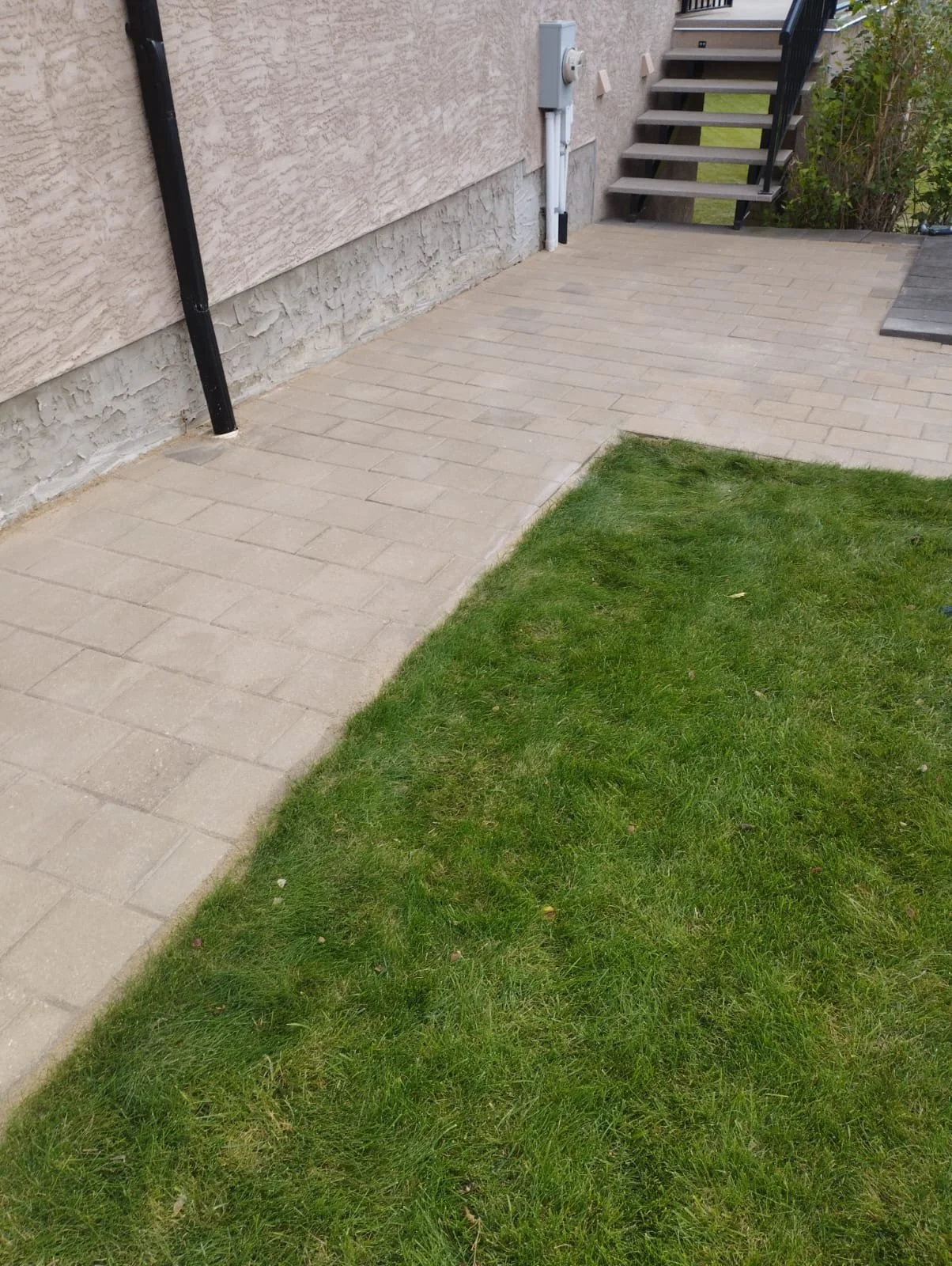 A small outdoor patio area with beige brick pavers, adjacent to a grassy lawn, with exterior walls, stairs, and utility fixtures visible.