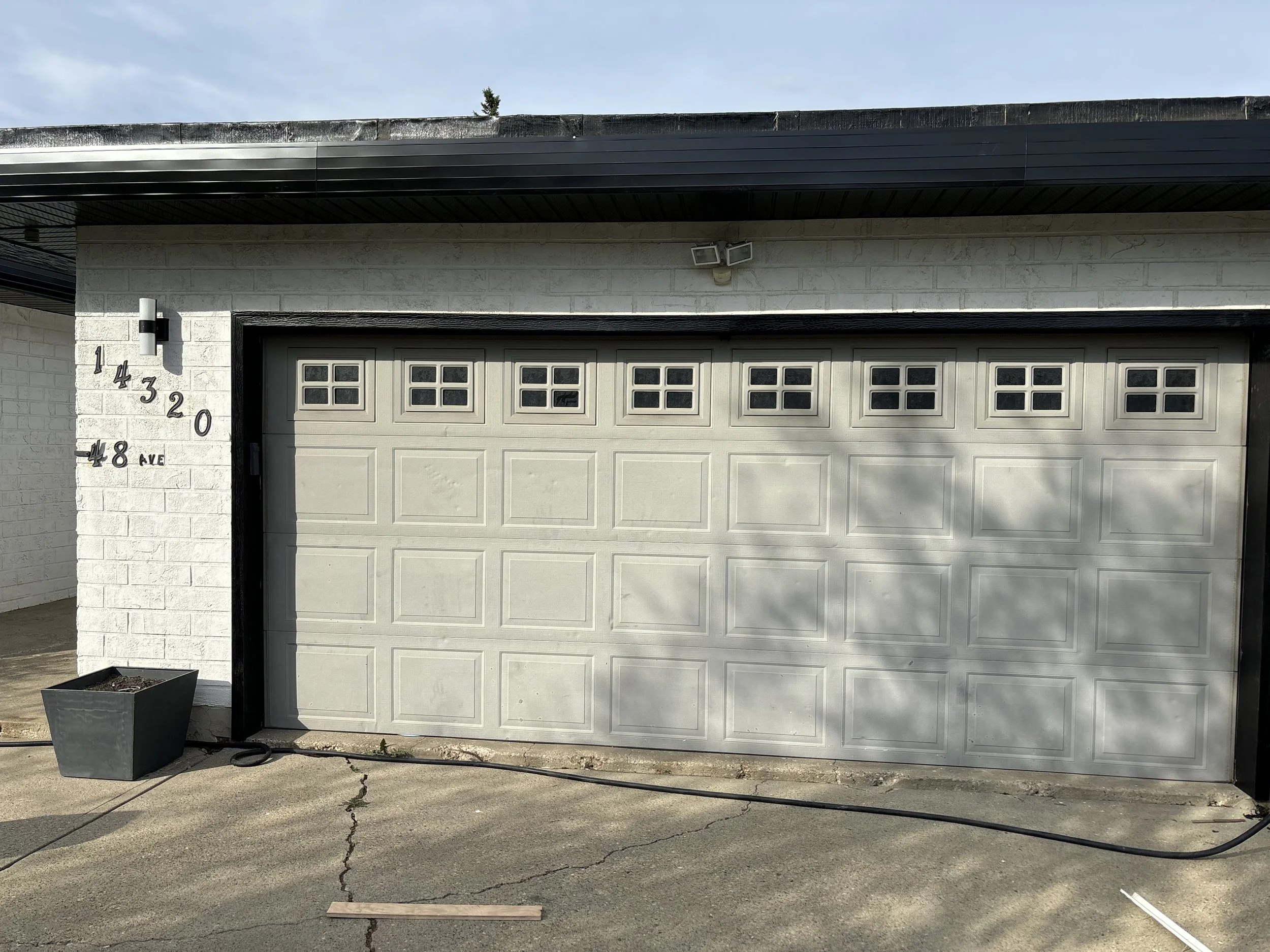 Gray garage door with small window panels at the top, part of a white brick building, house number 14320 and 48th Avenue on the left, black trim, paved driveway with some cracks and a black hose or cable on the ground.