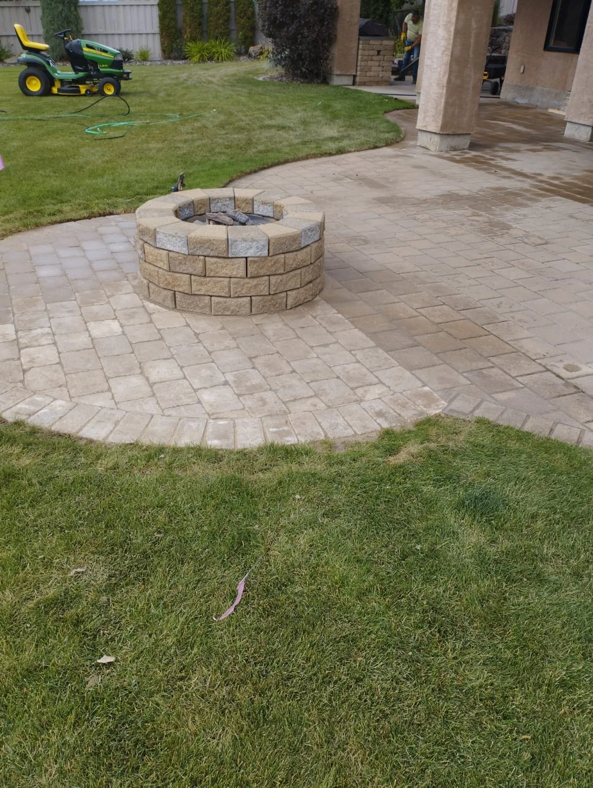 Backyard patio with a circular stone fire pit, brick pavers, a grassy area, a yellow riding lawn mower, and a person working in the background.