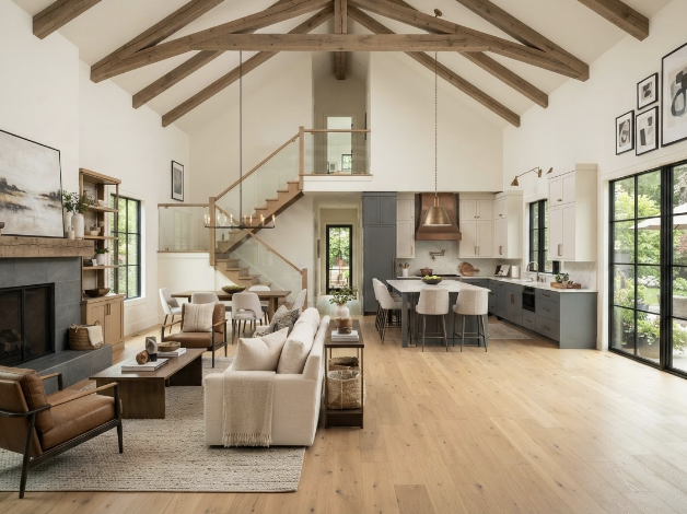 Open-concept living room and kitchen with vaulted ceiling and exposed wooden beams, large windows, light-colored walls, modern furniture, and a staircase leading to an upper level.