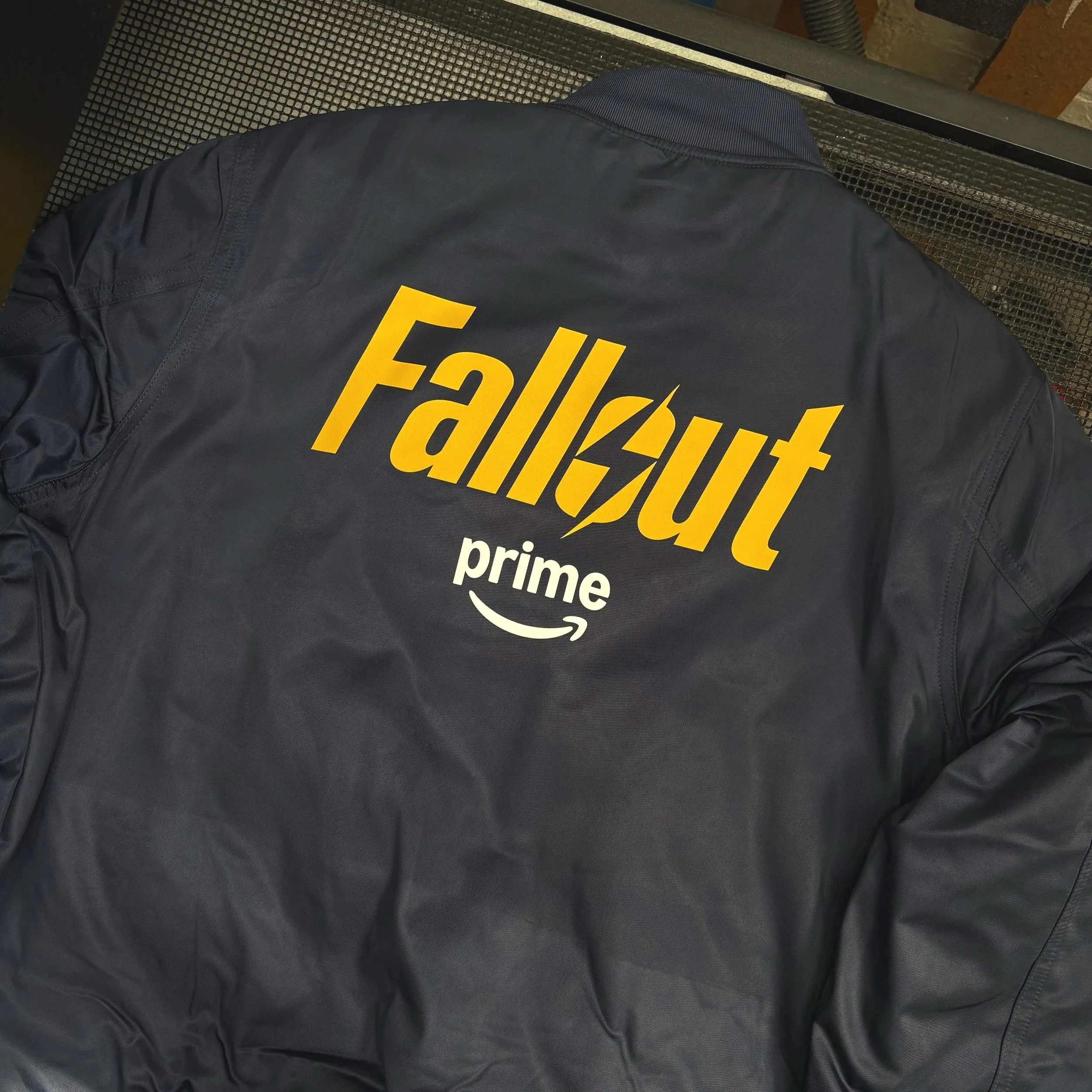 The image shows the back of a navy bomber jacket printed with a yellow logo of tv series Fall Out and an Amazon prime logo below it in white,