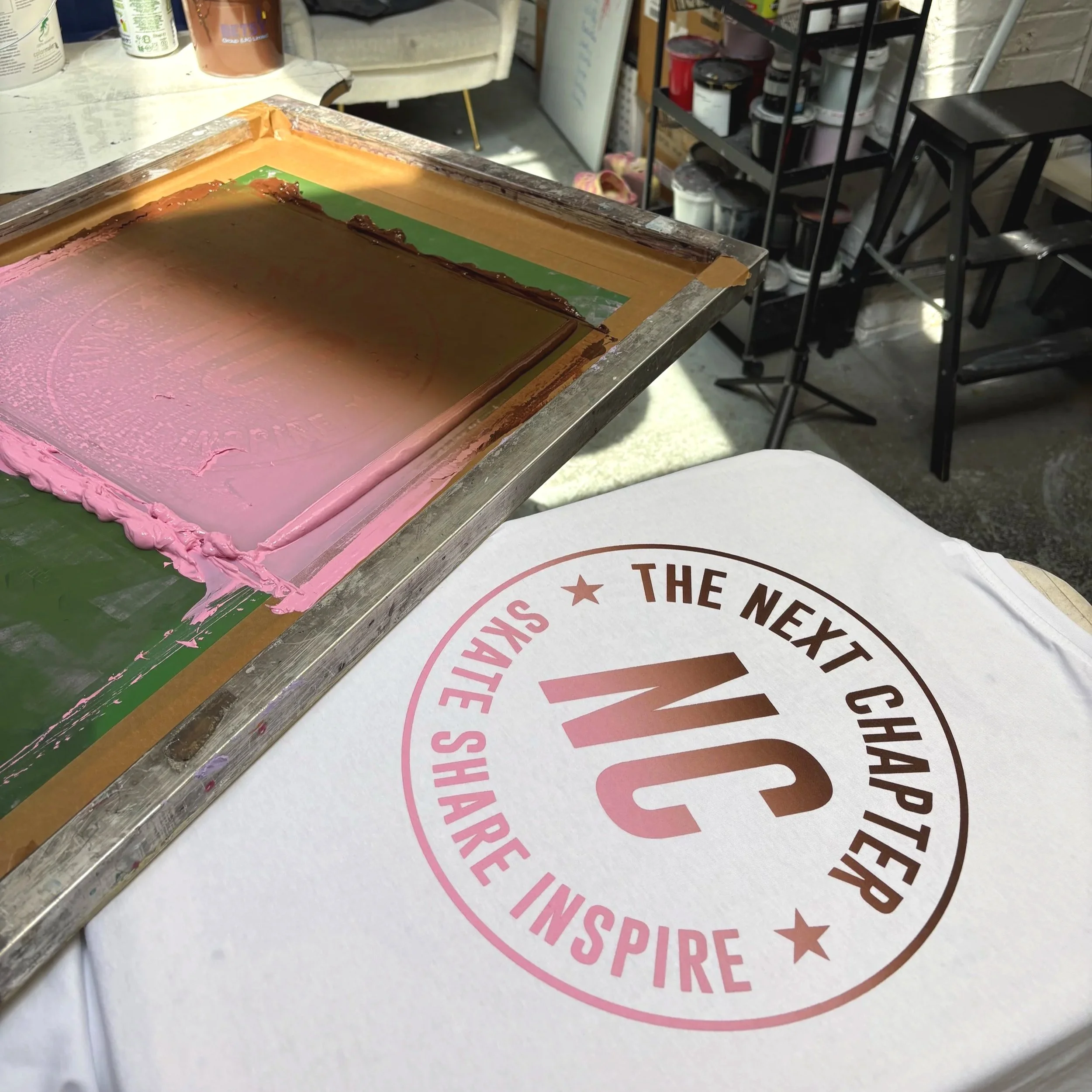 The image shows a screen printing screen with brown and pink inks blended horizontally, next to it is a white t-shirt, freshly screen printed using those inks. The print on the t-shirt reads NC - The Next Chapter, Skate, Share, Inspire.