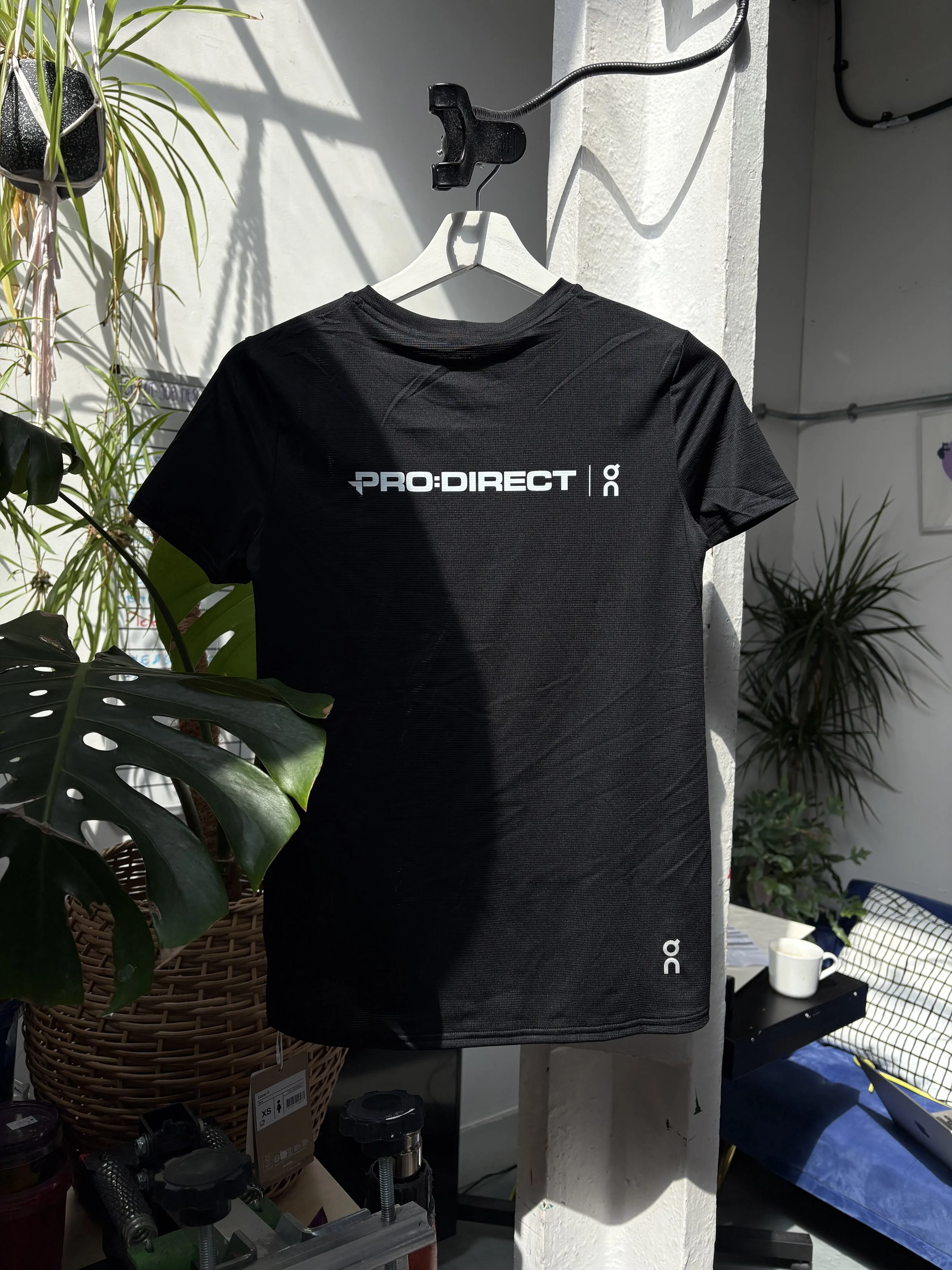 The Image shows a black T-shirt on a hanger, with plant leaves around it and white background. The t-shirt has a white print on it, which reads: Pro:Direct | On.