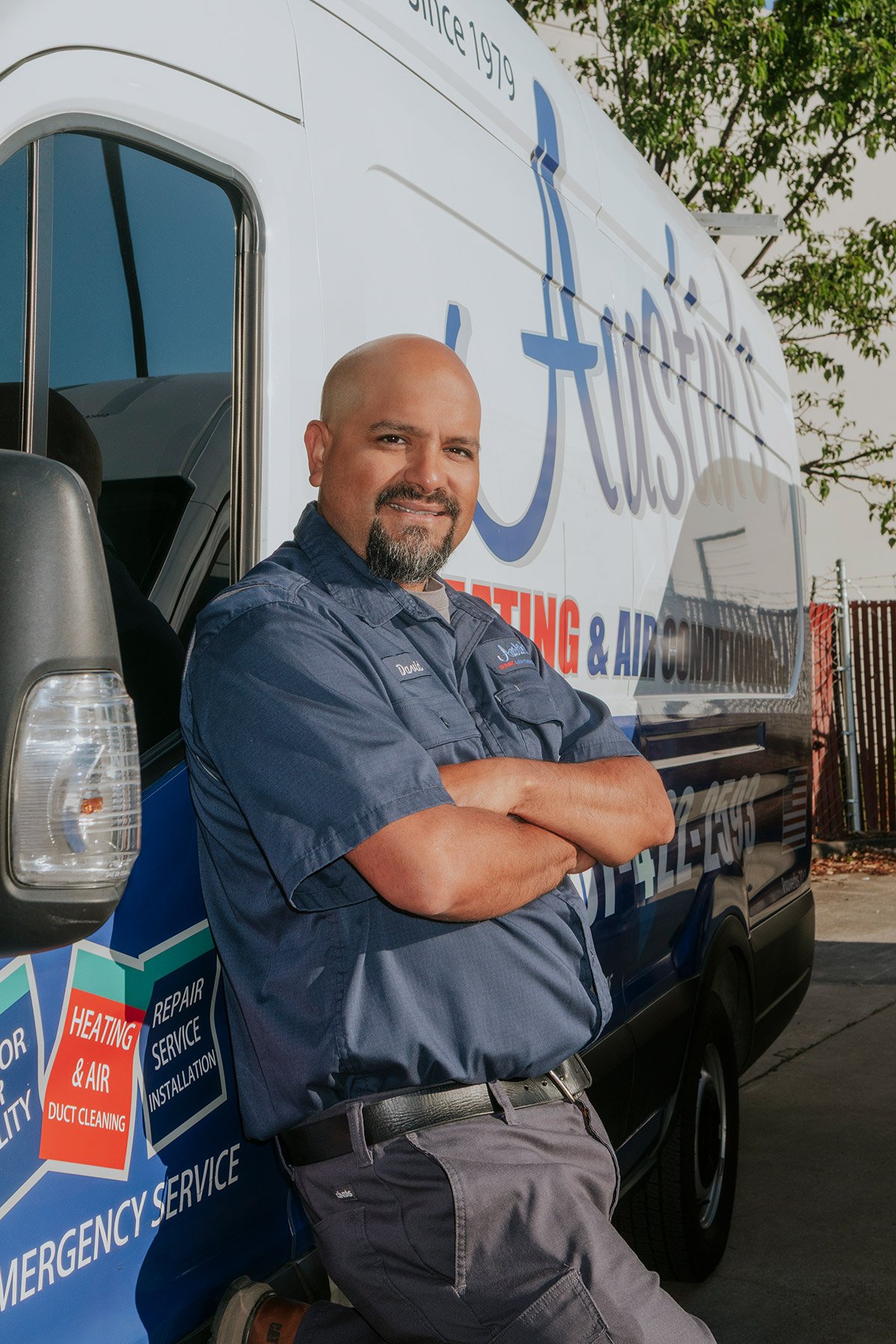 A man in a blue uniform leaning against a service van with company branding for heating, air conditioning, and ventilation repair and service.
