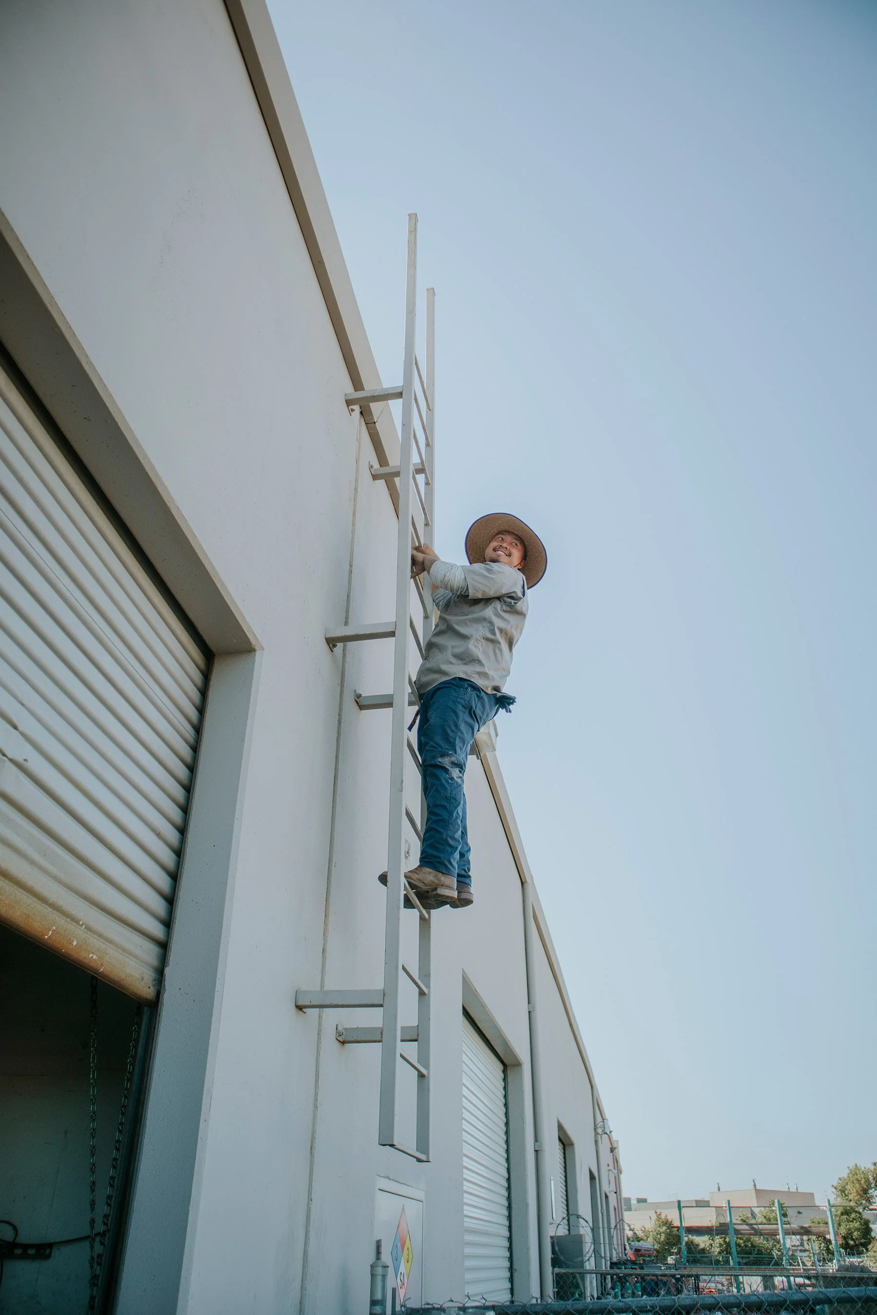 A person climbing an outdoor ladder attached to a building, wearing a wide-brimmed hat and smiling up at the sky.