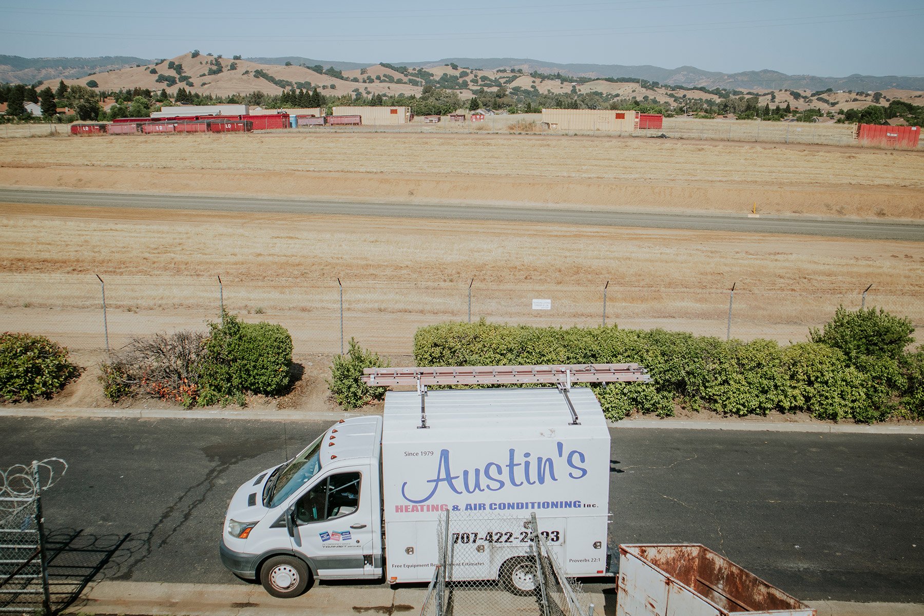 White service van with company name 'Austin's Heating & Air Conditioning Inc.' parked on asphalt, with bushes and a chain-link fence in the foreground, and a large open area with a railroad track and hills in the background.