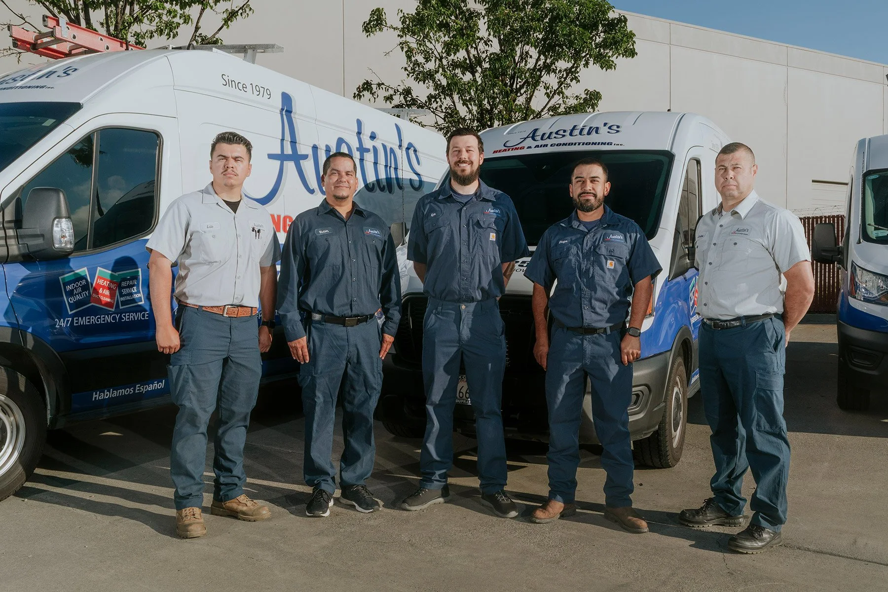 Five HVAC technicians standing in front of service vans with the Austins logo, outdoors on a sunny day.
