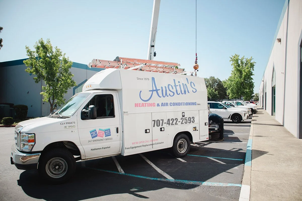 White service van for Austin's Heating & Air Conditioning parked in a lot, with a worker accessing the back, and other parked cars nearby, under a clear blue sky.