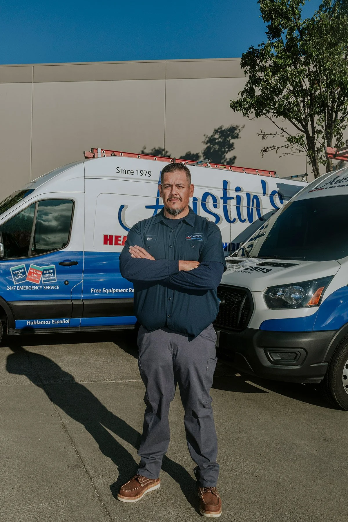 A man standing with arms crossed in front of company service vans outdoors on a sunny day.