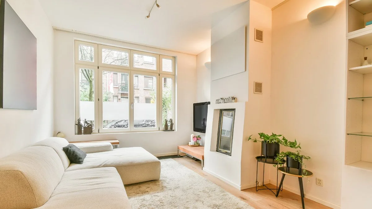 Bright living room with beige L-shaped sofa, large window, white walls, TV, potted plants, and bookshelf.