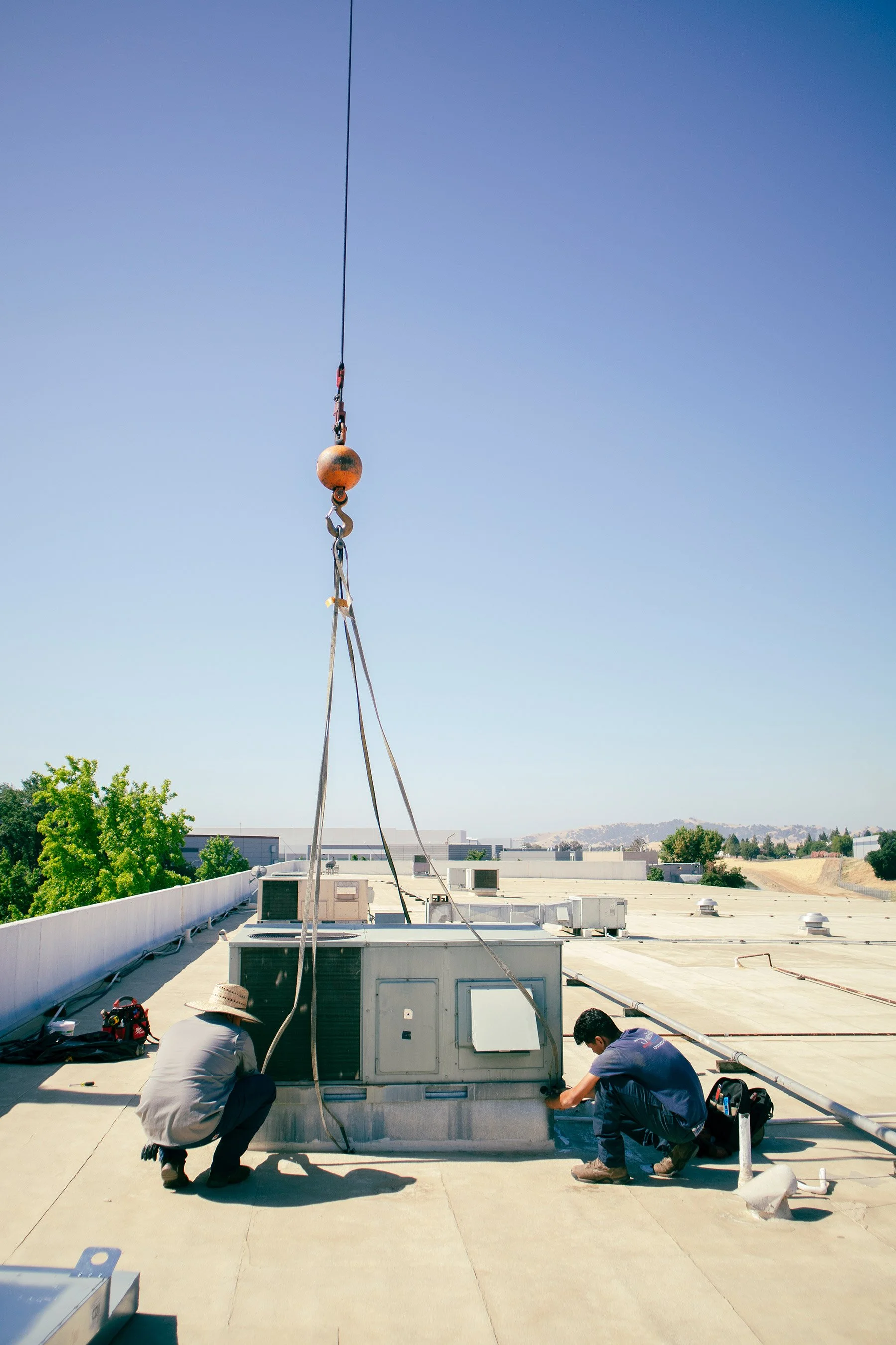 Two technicians working on an air conditioning unit on a building rooftop, with a crane hook above them.