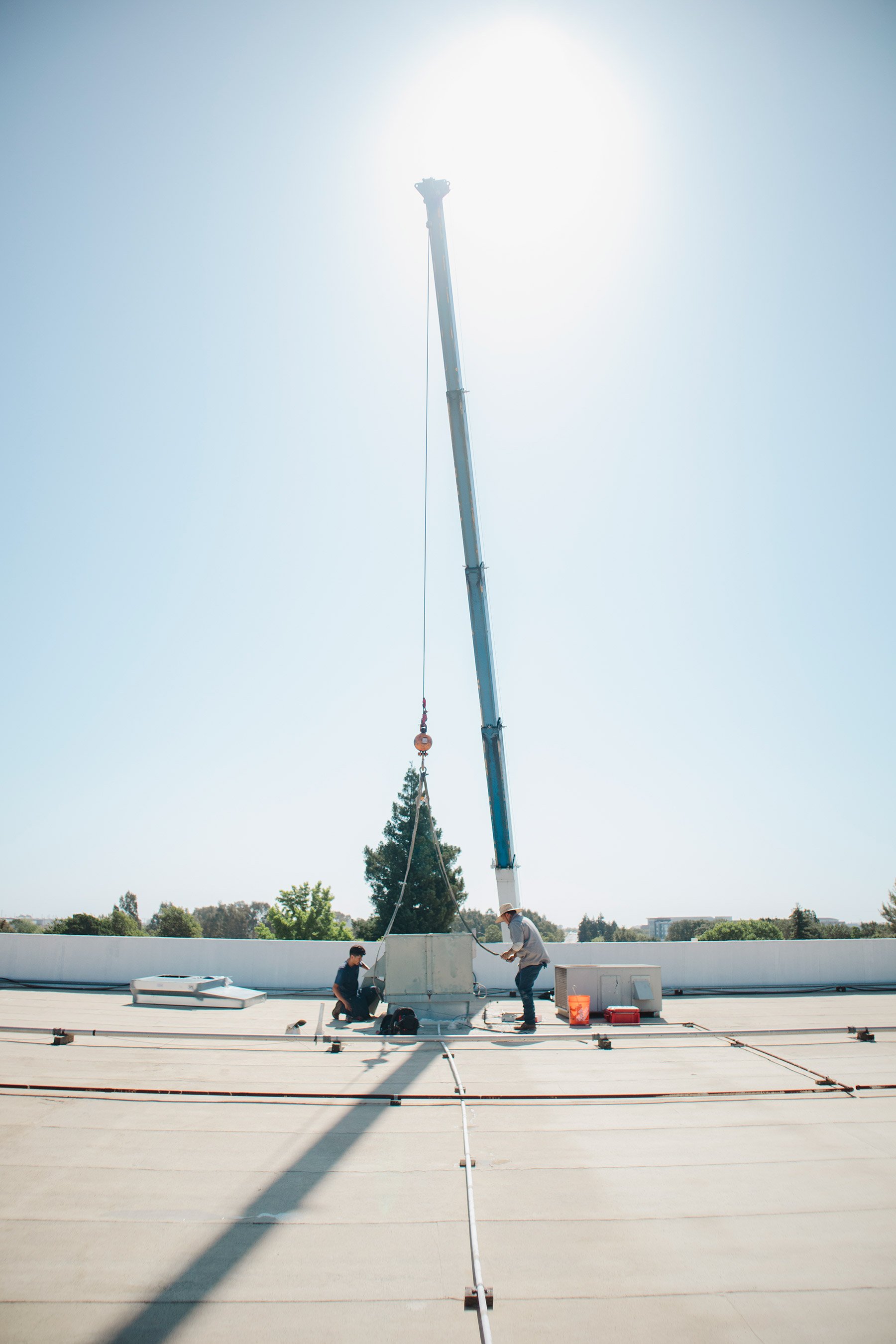 Two workers inspecting a large rooftop air conditioning unit, with a crane overhead in bright sunlight.