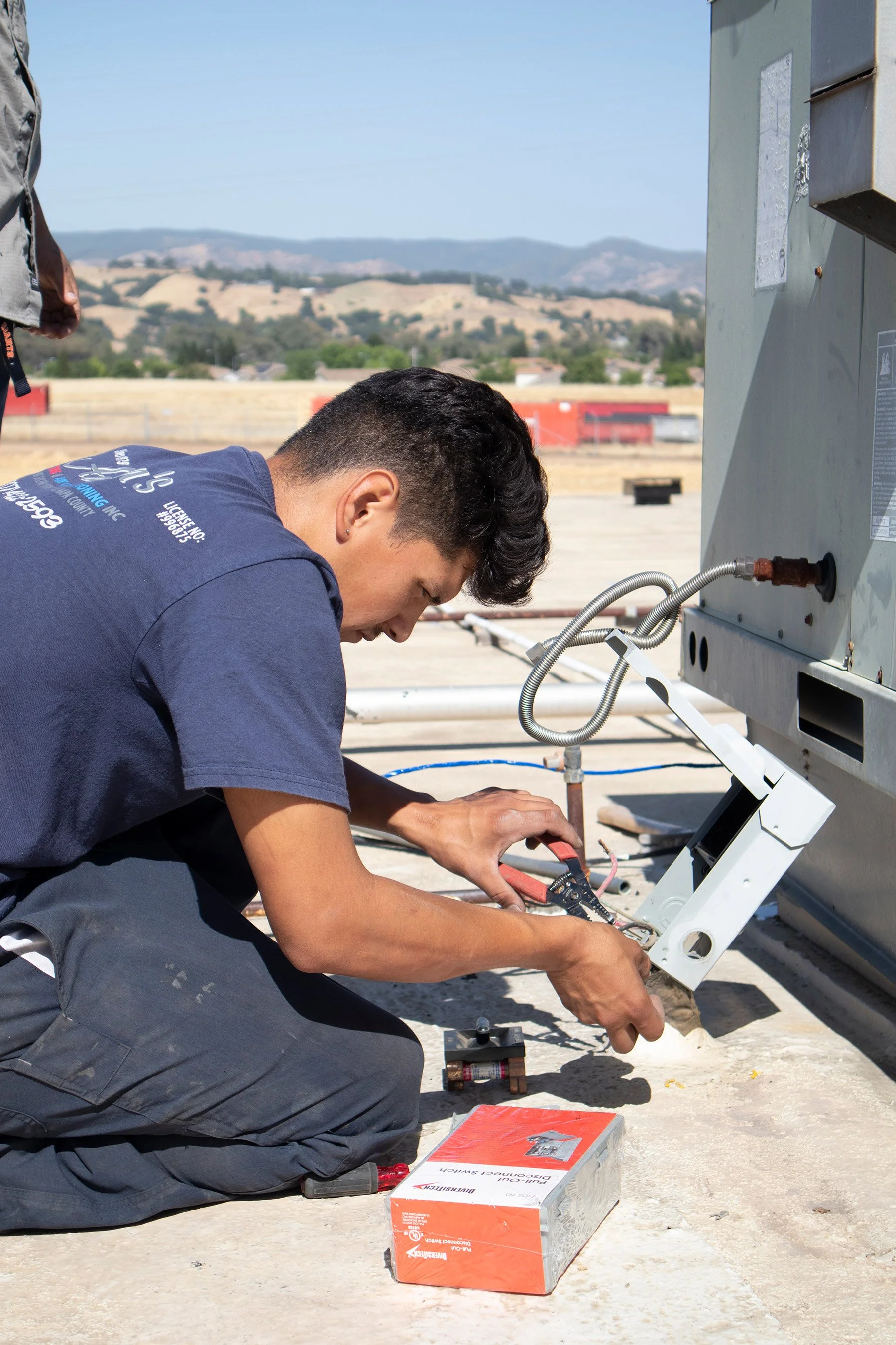 A technician kneeling on a rooftop using a multimeter to test electrical connections on HVAC equipment with a scenic view of hills in the background.