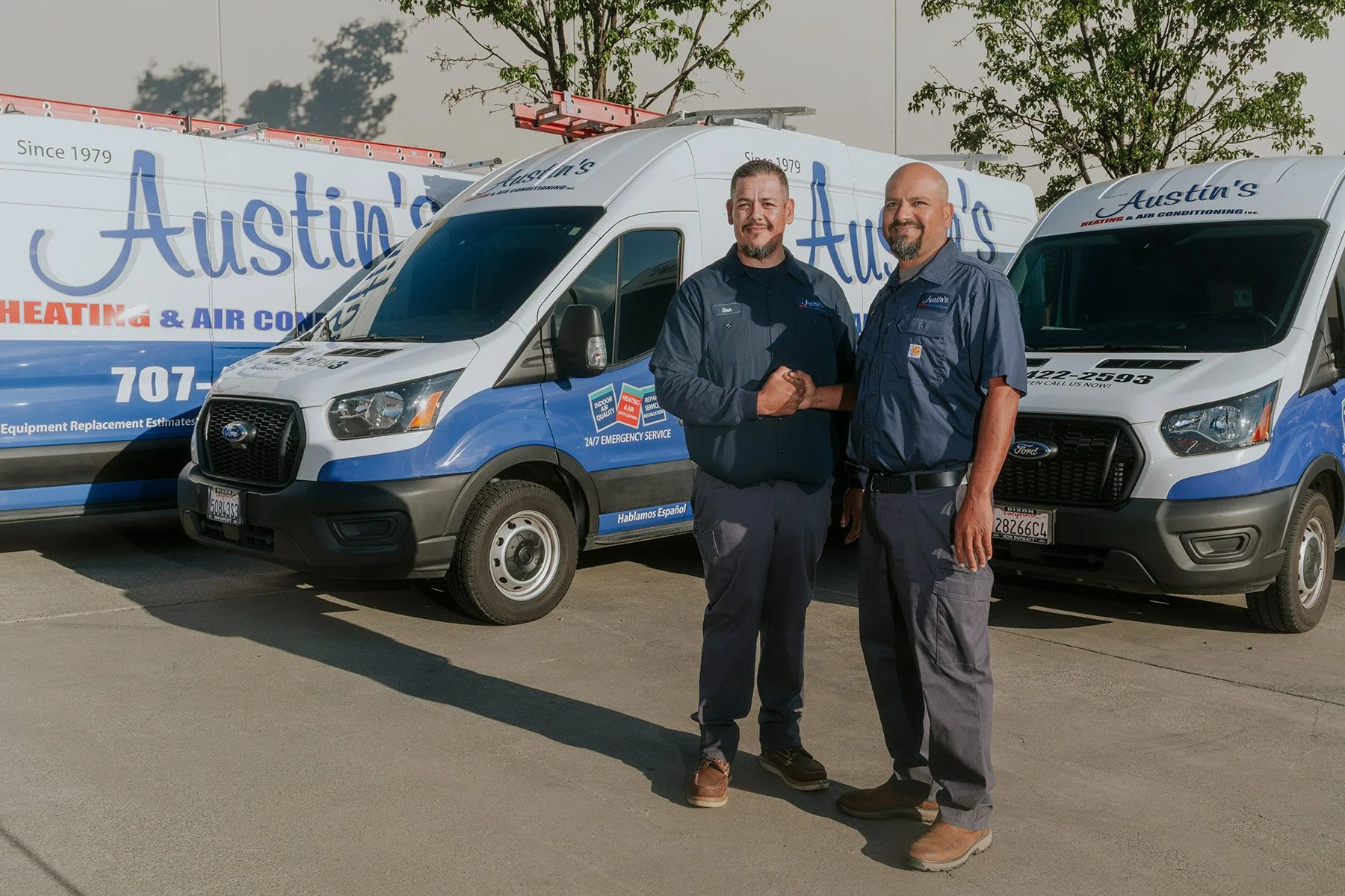 Two men in work uniforms shaking hands in front of company service vans for Austin's Heating & Air Conditioning, parked outdoors on a sunny day.