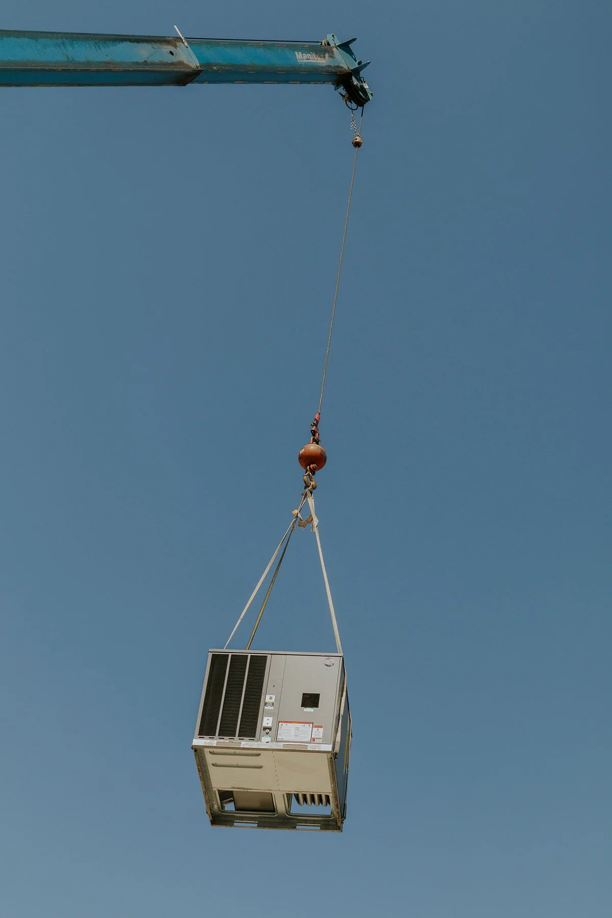 A crane lifting a large air conditioning unit against a clear blue sky.