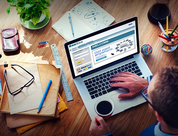 A person working on a laptop at a wooden desk surrounded by notebooks, folders, a ruler, a pen, glasses, a cup of coffee, a potted plant, and colorful office supplies.