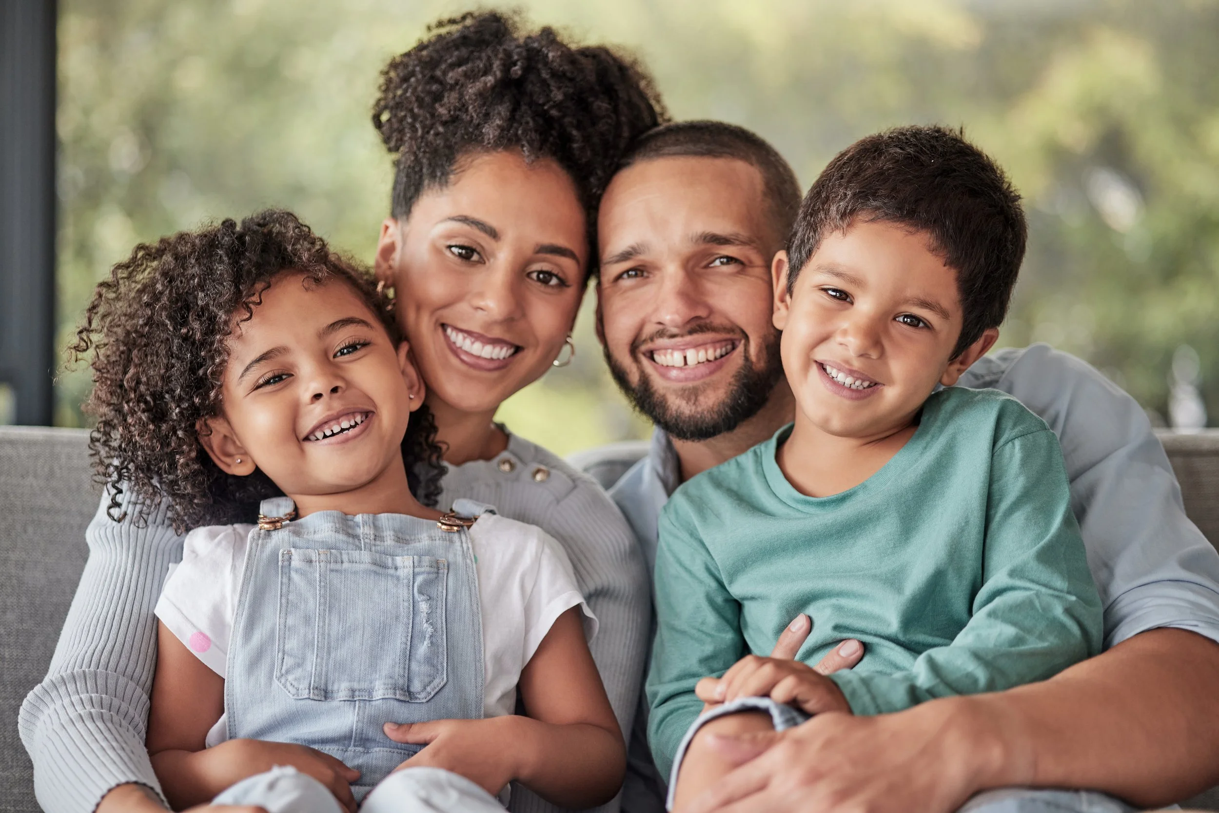 A family of four smiling and sitting close together on a couch.