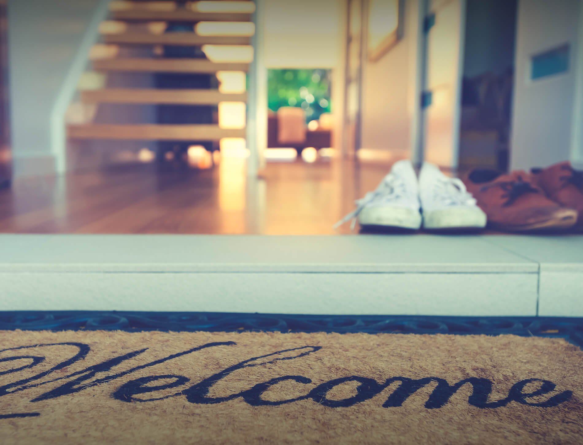 View from outside a door showing a welcome mat with the word 'Welcome', a pair of white sneakers, and brown shoes, with a staircase and door leading outside in the background.