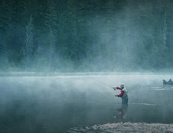 A person fishing in a misty lake with a dense forest in the background and a boat in the distance.