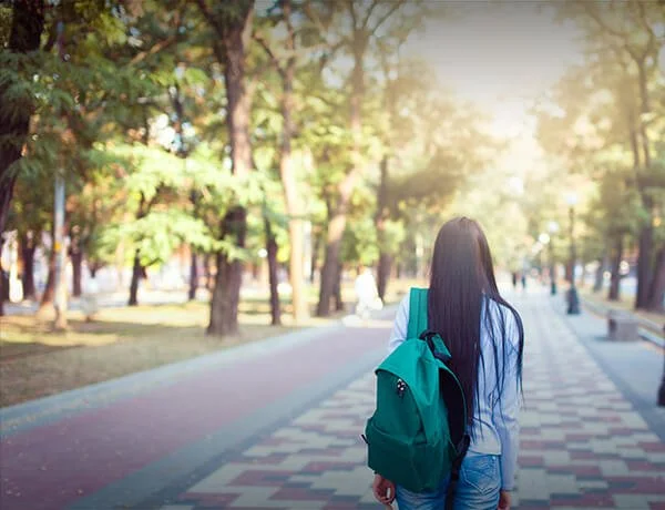 Young woman walking in a park with a teal backpack, trees and sunlight in the background.